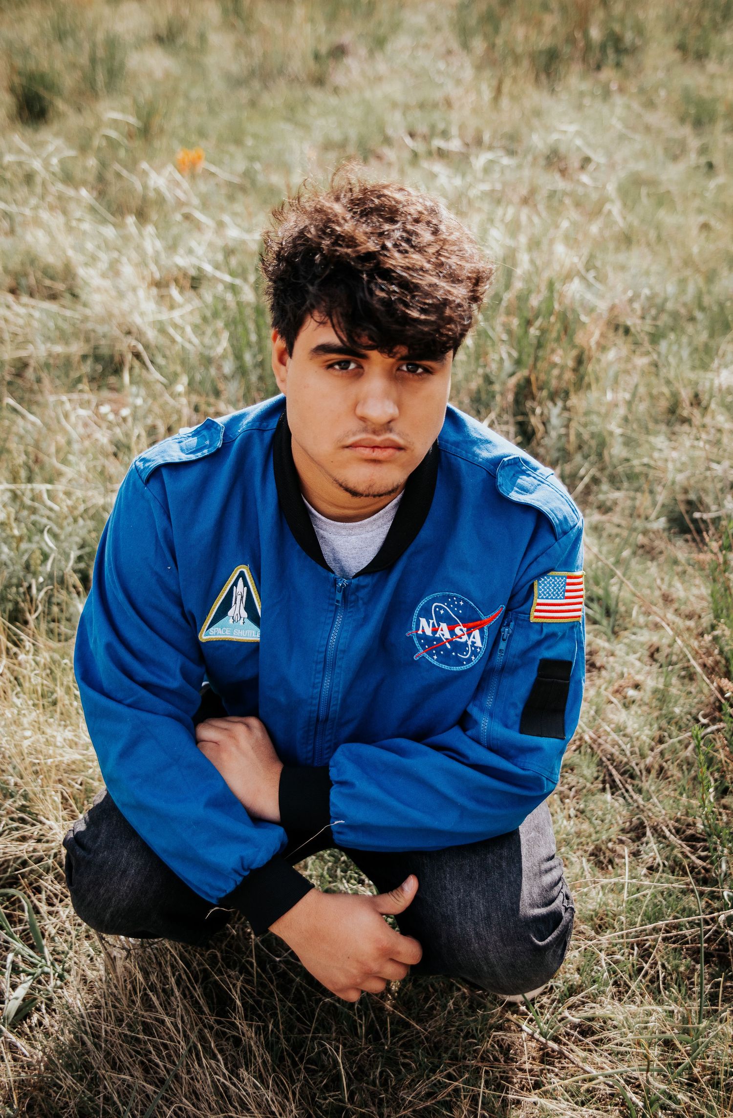 High school senior in official NASA jacket crouching in field of wild grass at Palmer Park in Colorado Springs