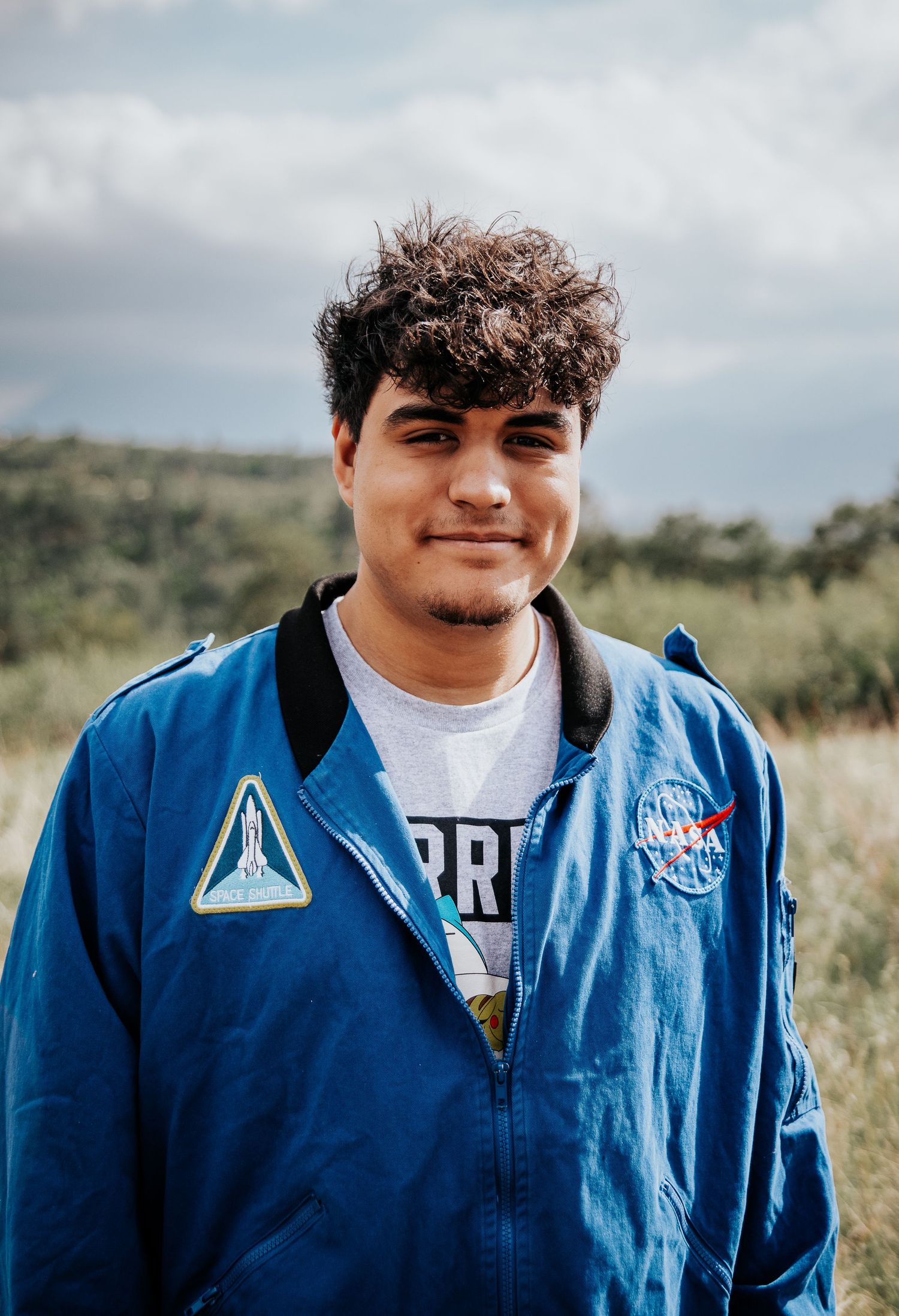 High school senior in blue NASA jacket smiles while posing outdoors on a sunny day at Palmer Park in Colorado Springs