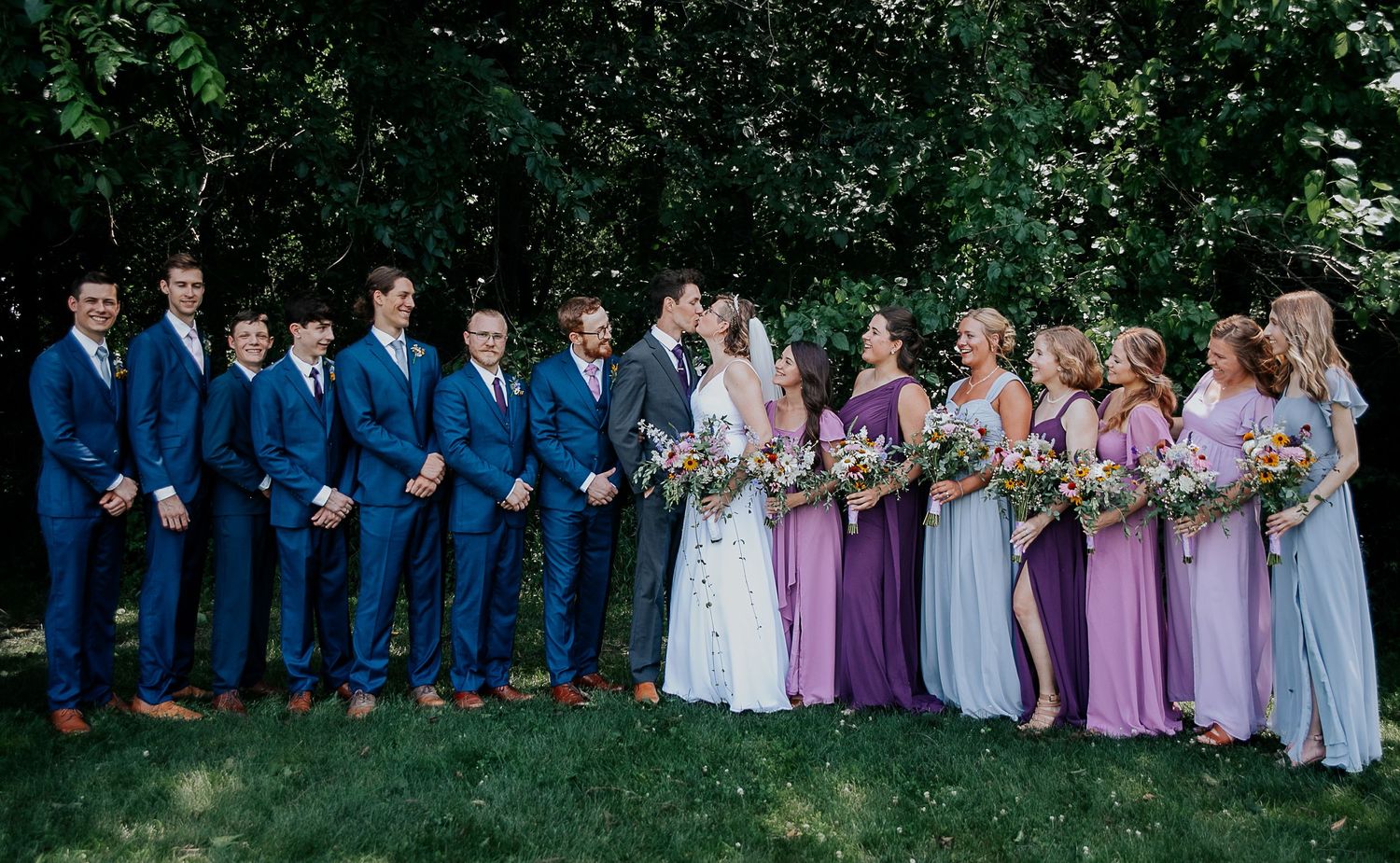 Creative wedding photo shows couple kissing while bridal party poses playfully in background.