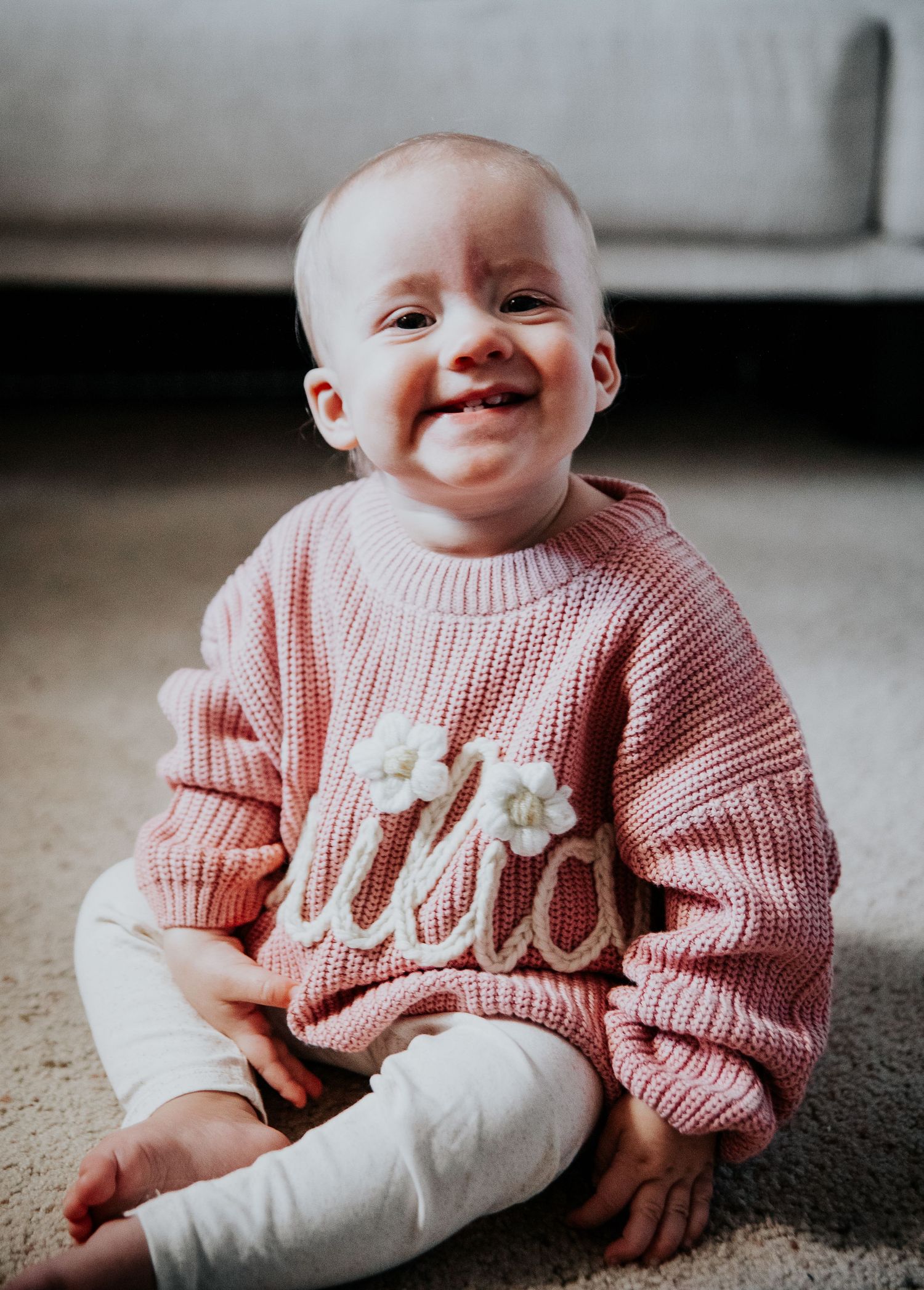 Baby wearing pink knit sweater sits on carpet showing happy expressions.