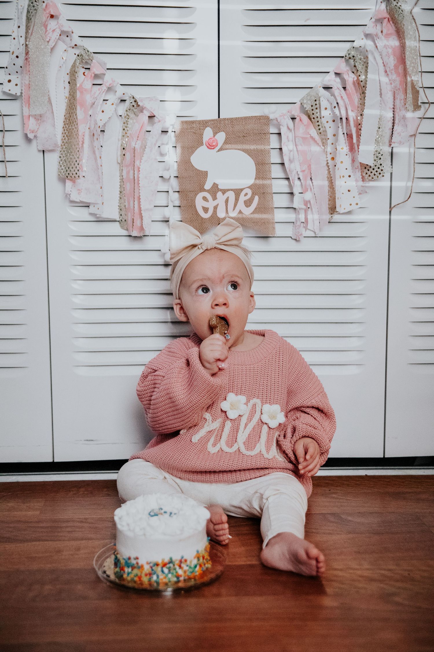 Baby in pink sweater explores white frosted cake during first birthday photoshoot.