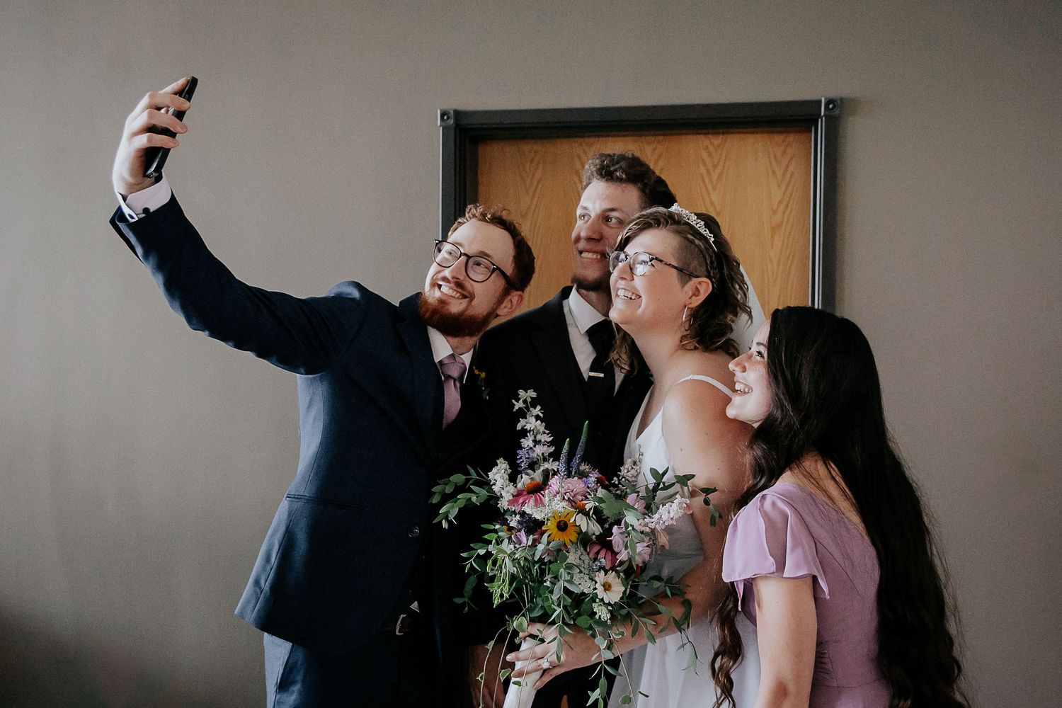 Wedding party takes a group selfie with a smartphone while holding a floral bouquet.