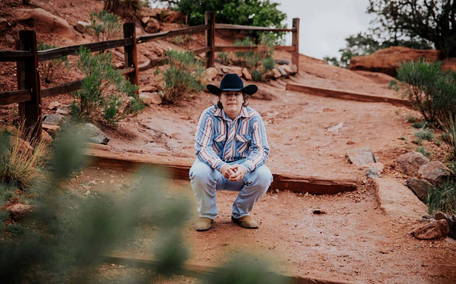 A high school senior in a striped western shirt and cowboy hat sits on red rocks at Garden of the Gods in Colorado Springs