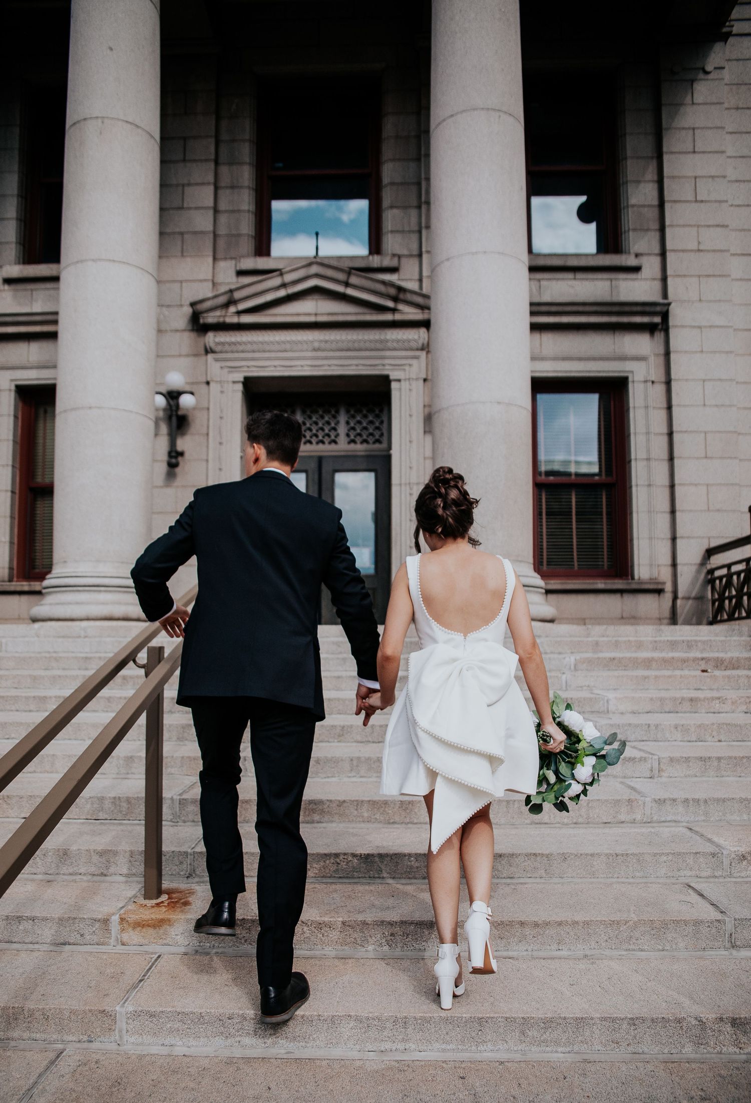 Wedding couple and guests ascending stone steps of historic building with white columns in progressive sequence.
