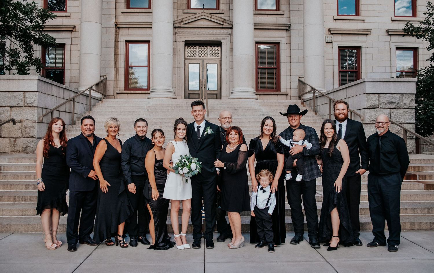 Panoramic wedding party group photo against classic stone building facade.