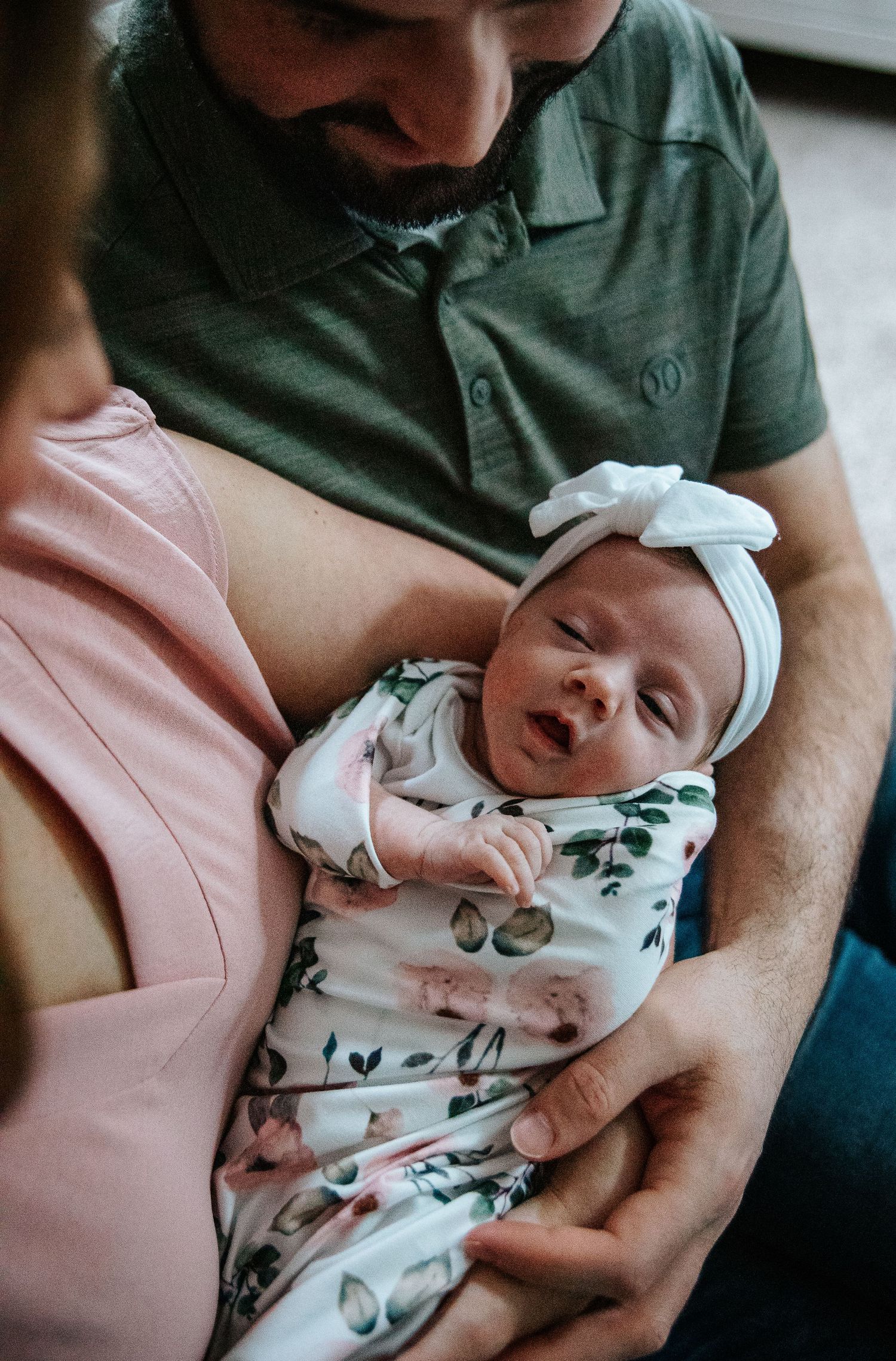 Newborn baby wearing floral outfit and white bow headband rests peacefully in parents' arms.
