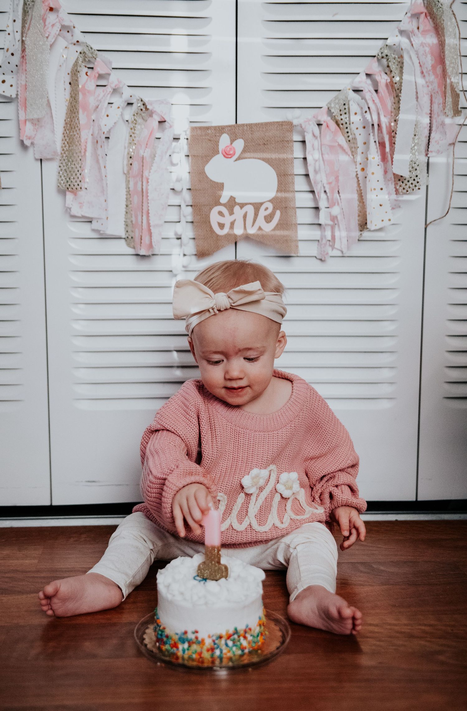 Baby in pink knitted sweater sits on wooden floor with white birthday cake and sparkly banner.