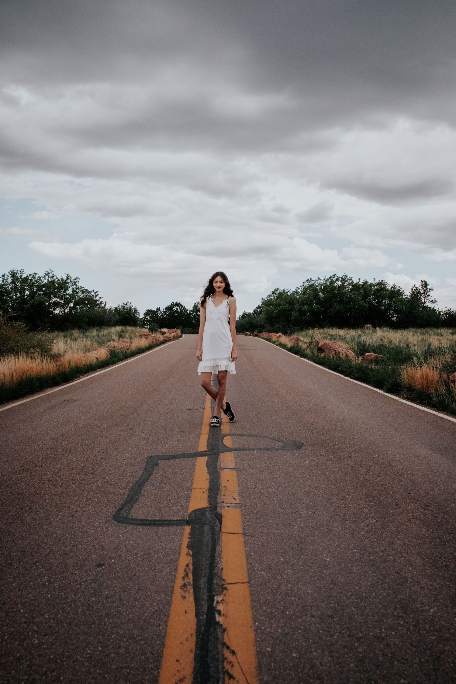Figure in white dress stands on empty road with dramatic cloudy sky above.