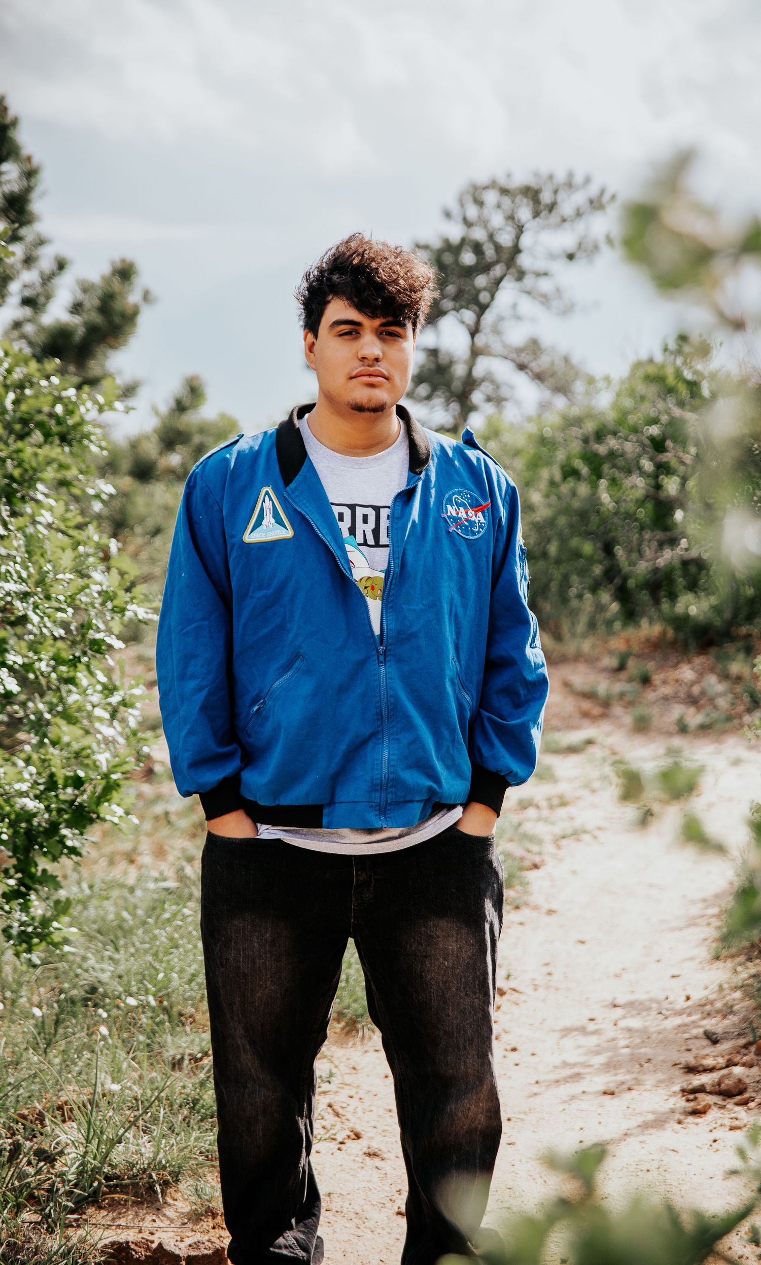 High school senior in blue NASA jacket poses on rustic mountain path amid desert vegetation at Palmer Park in Colorado Springs