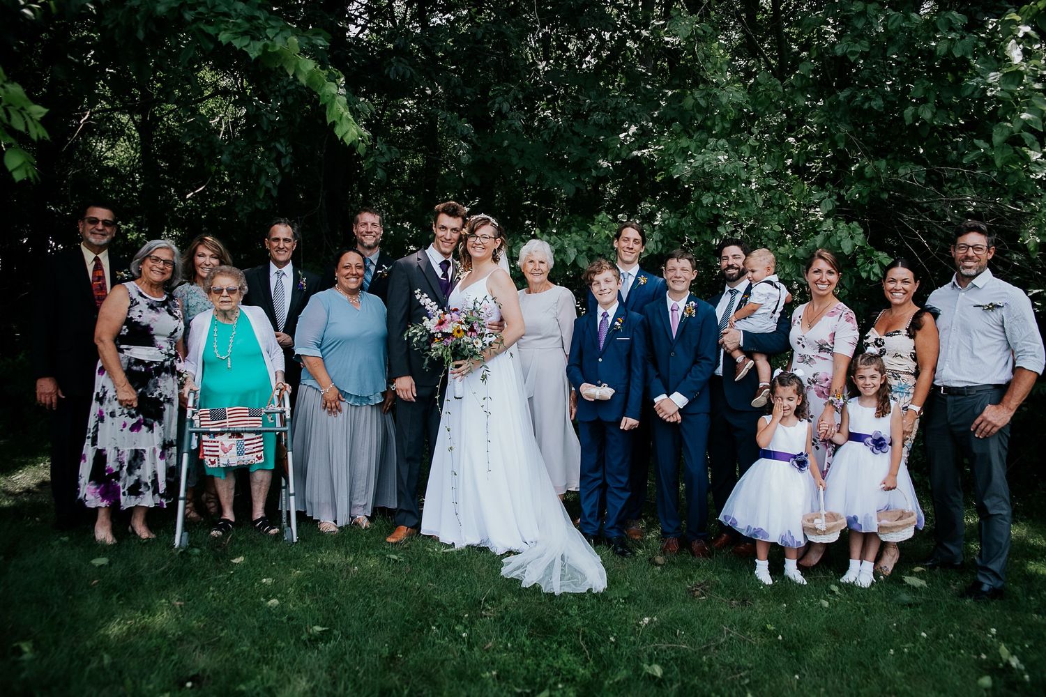 Large wedding party poses together outdoors in front of green trees and foliage.