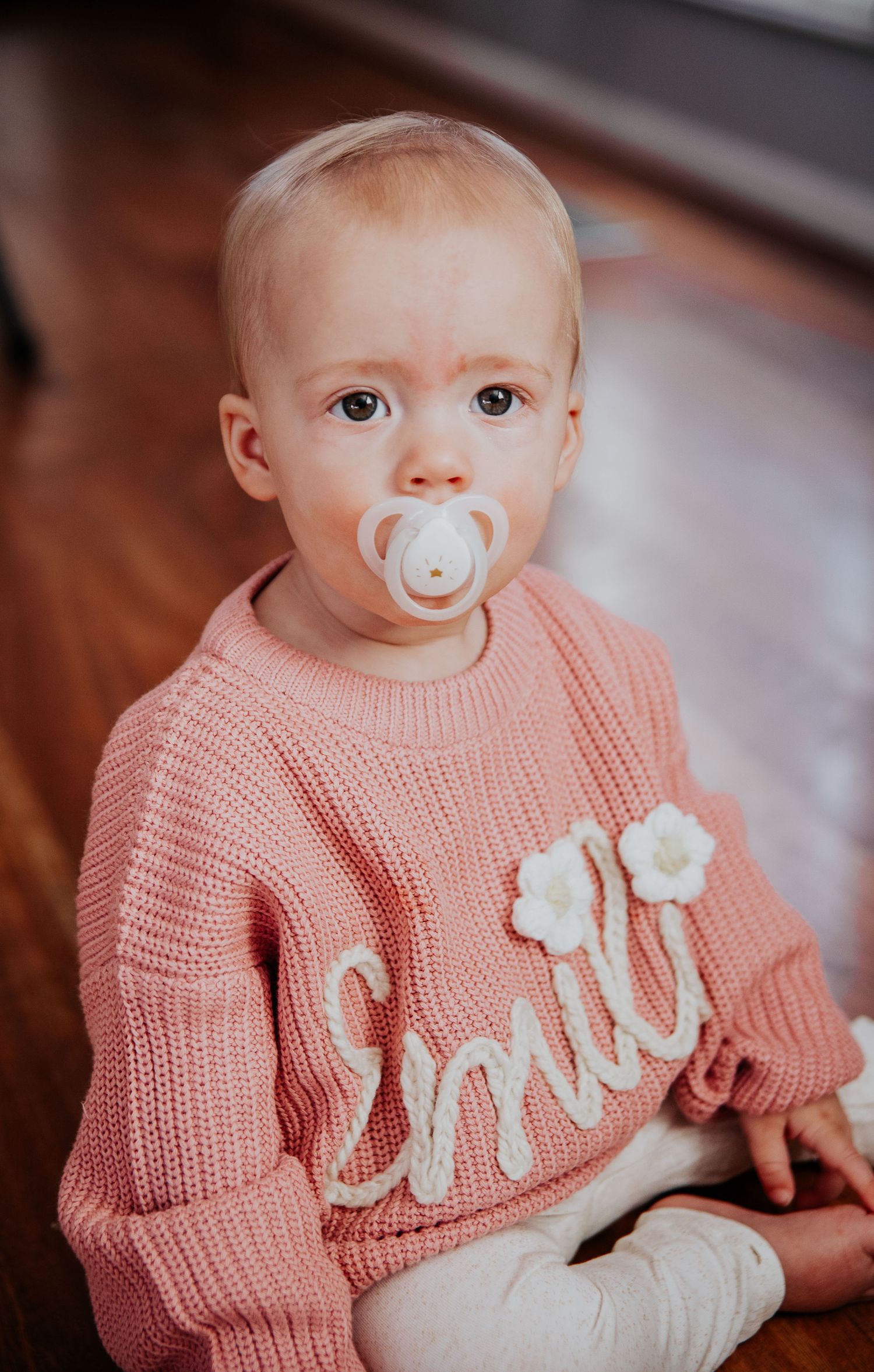 Baby wearing pink knit sweater with 'smile' text and white pacifier.