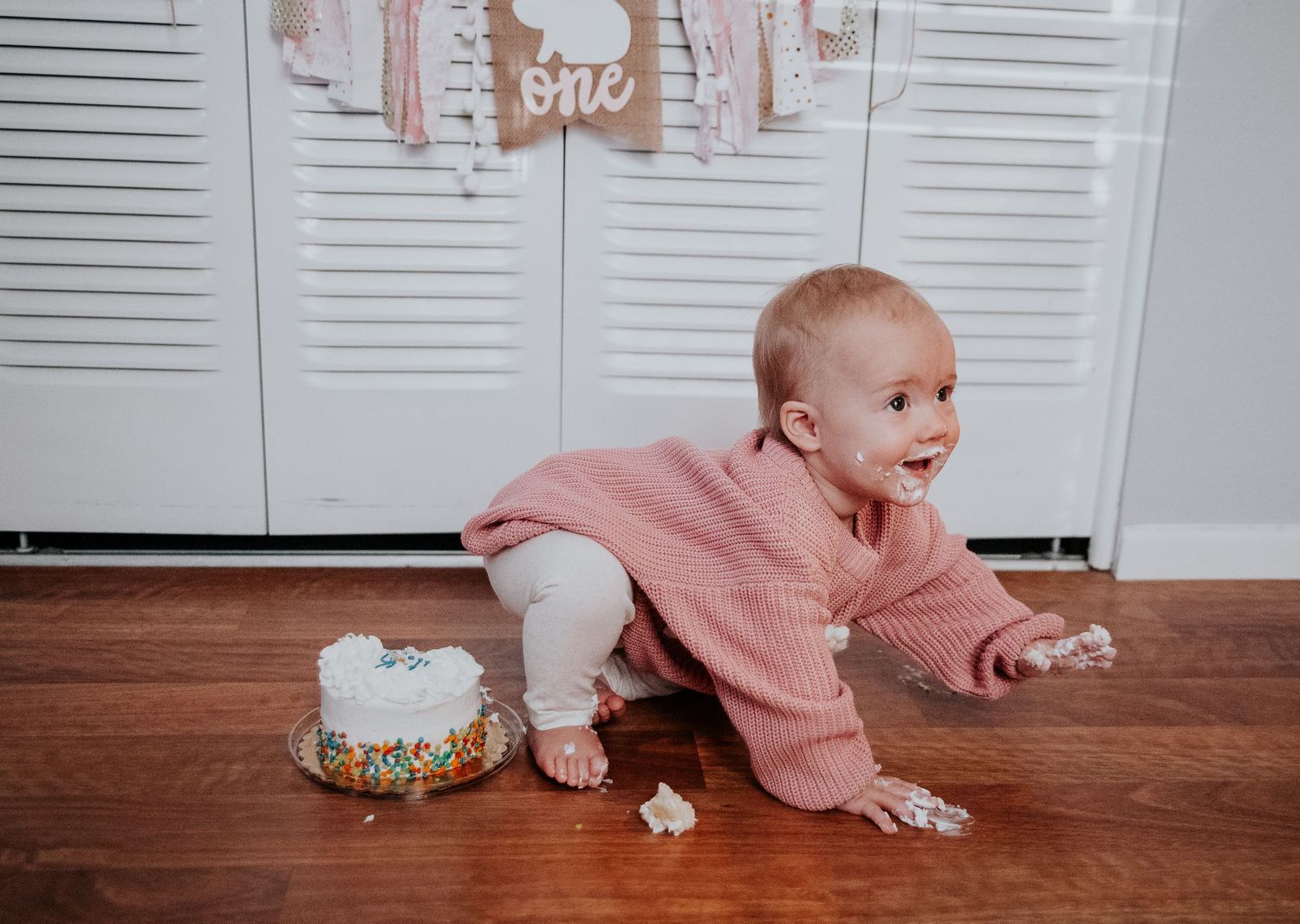 A cake smash photo session with pink decor and a crawling baby on hardwood flooring.