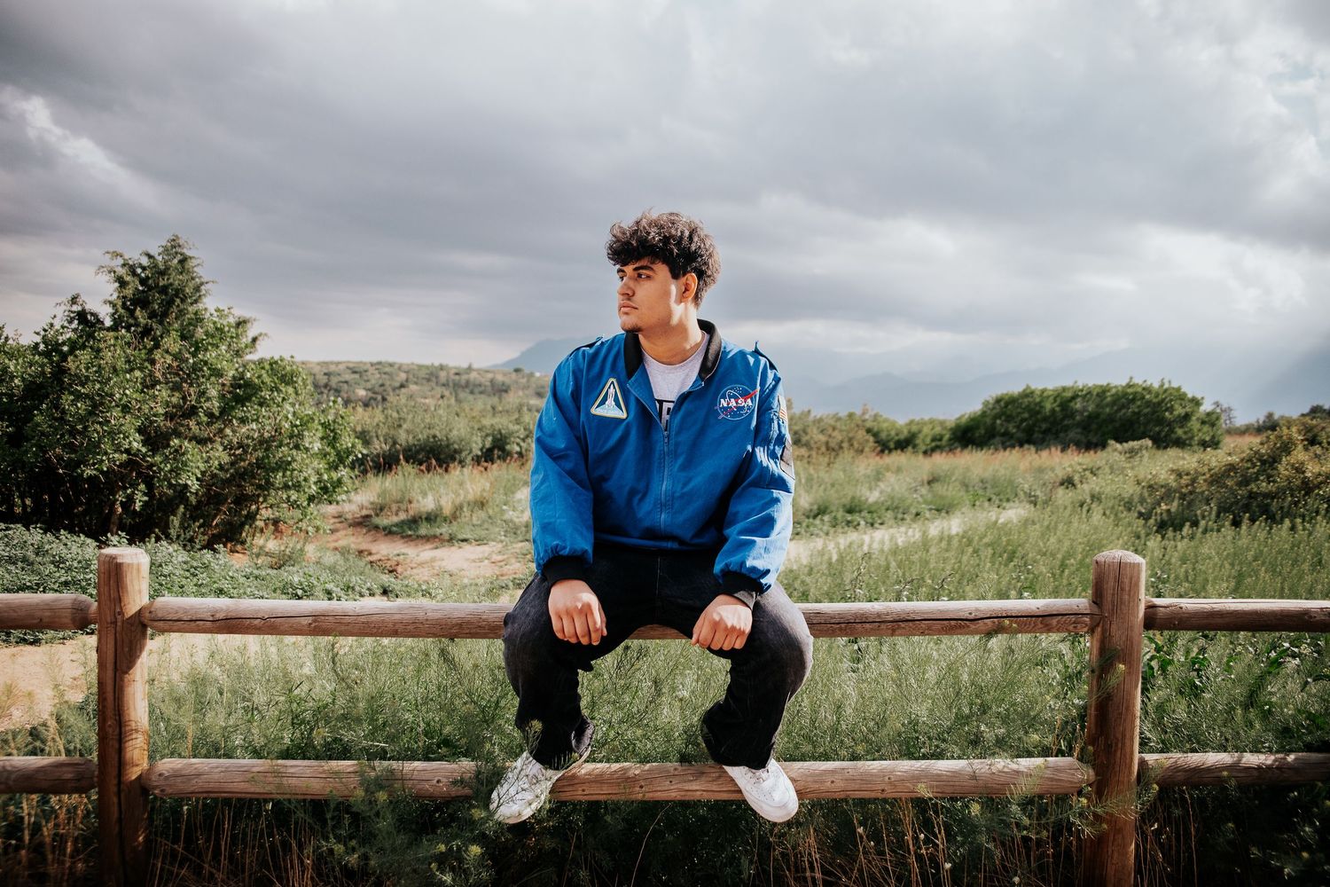 High school senior perched on wooden fence rail wearing blue jacket against cloudy sky backdrop at Palmer Park in Colorado Springs