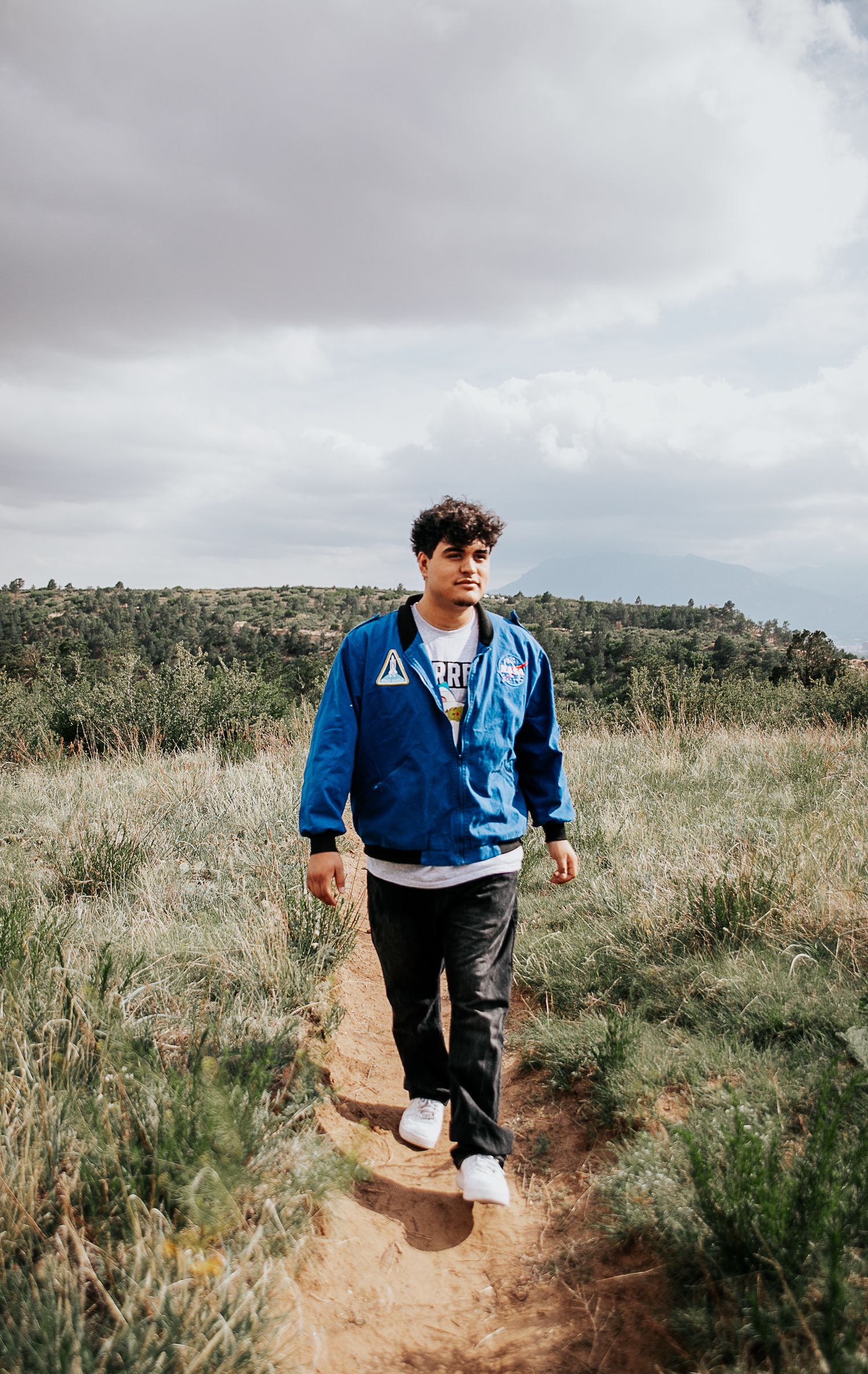 High school senior wearing a blue NASA flight jacket walks along a dirt hiking trail with mountains in the background at Palmer Park in CO