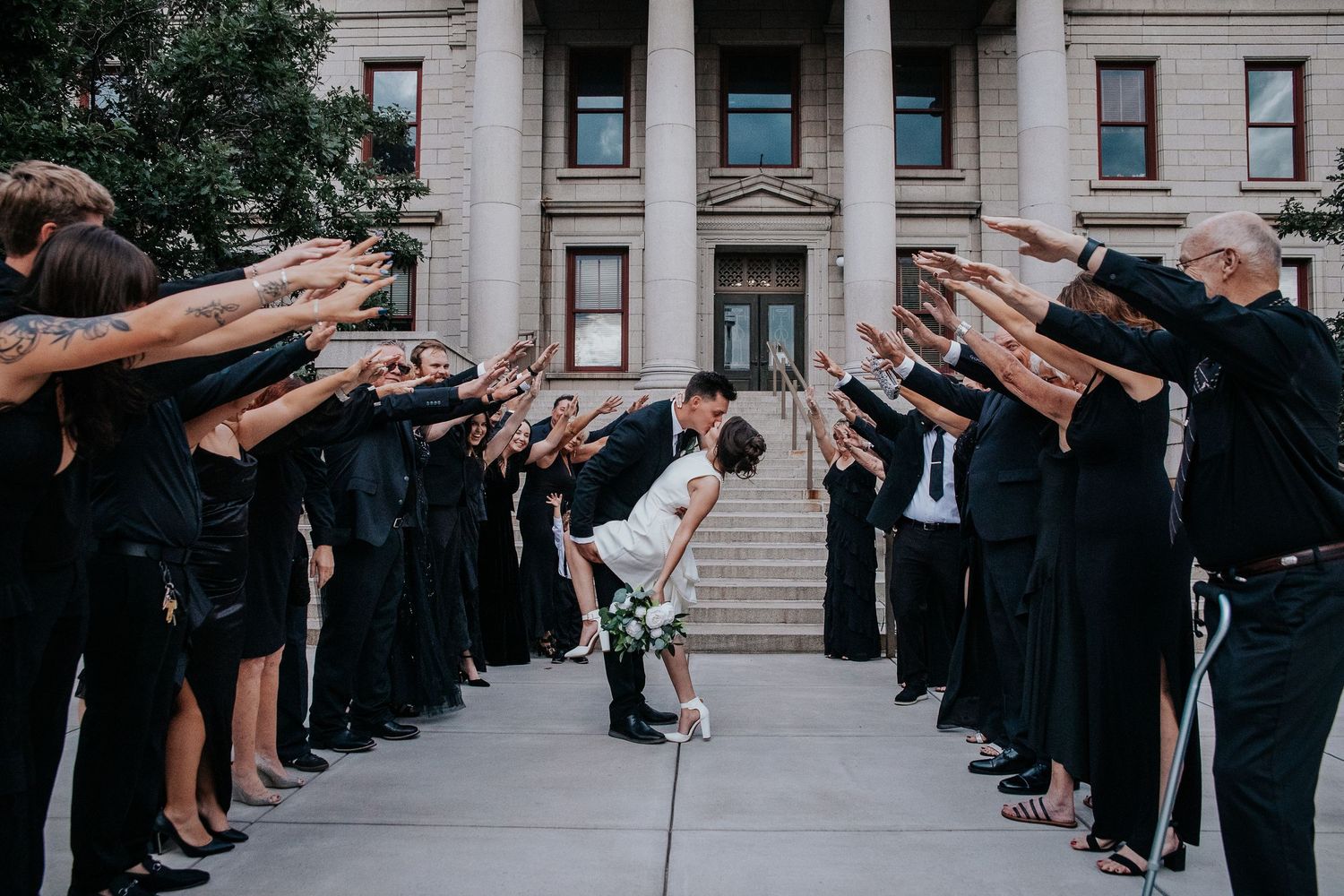 Wedding guests form a celebratory tunnel with raised arms outside a grand building.