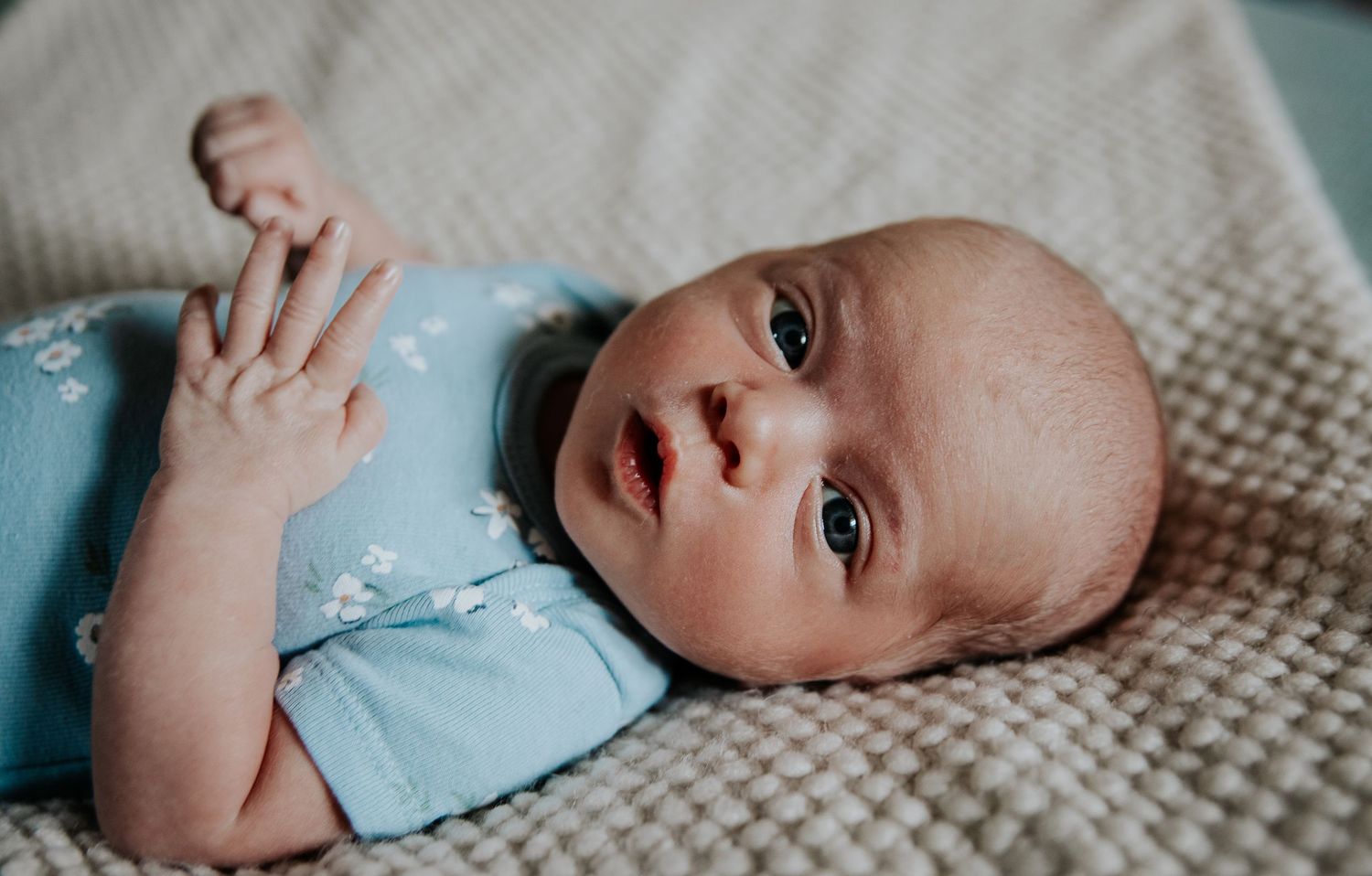Infant in light blue outfit lying on textured beige blanket.