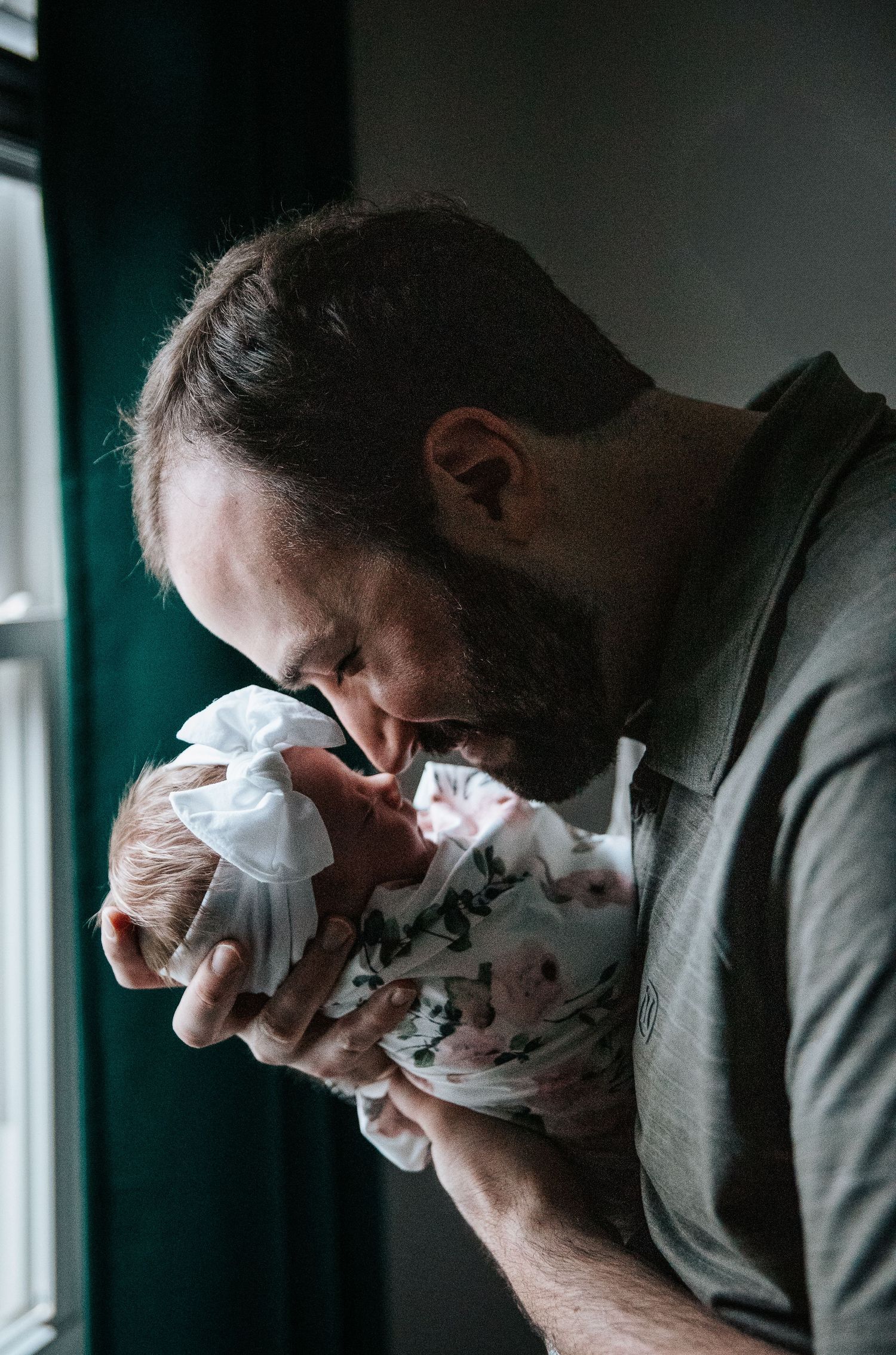 Parent tenderly holding and kissing newborn baby by window in moody lighting.