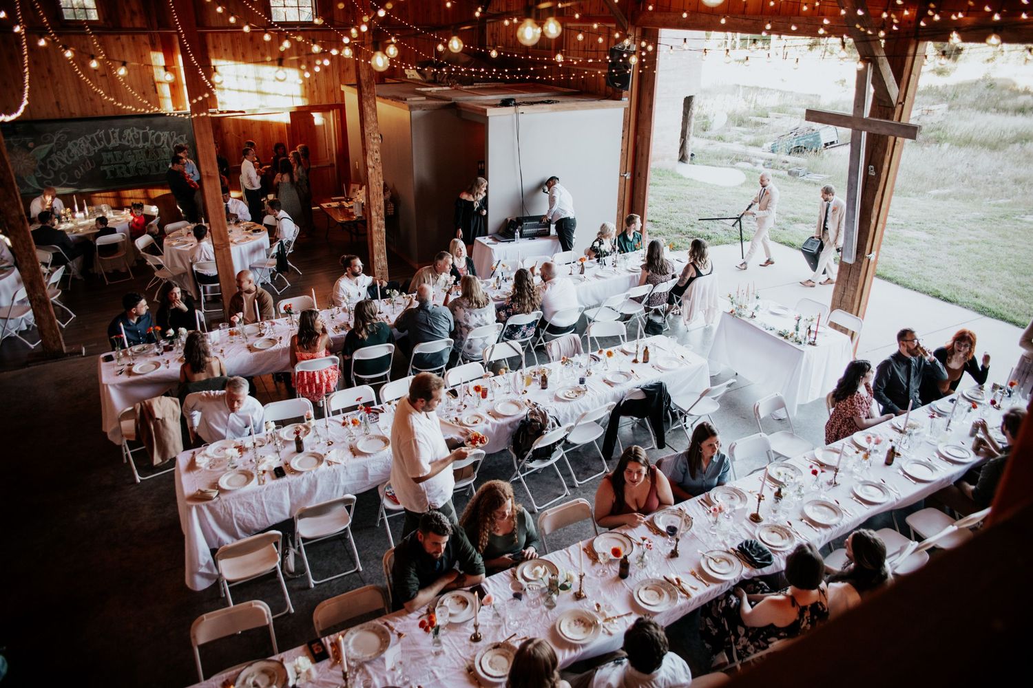 Rustic barn wedding reception with long tables set for dinner under string lights and wooden beams.