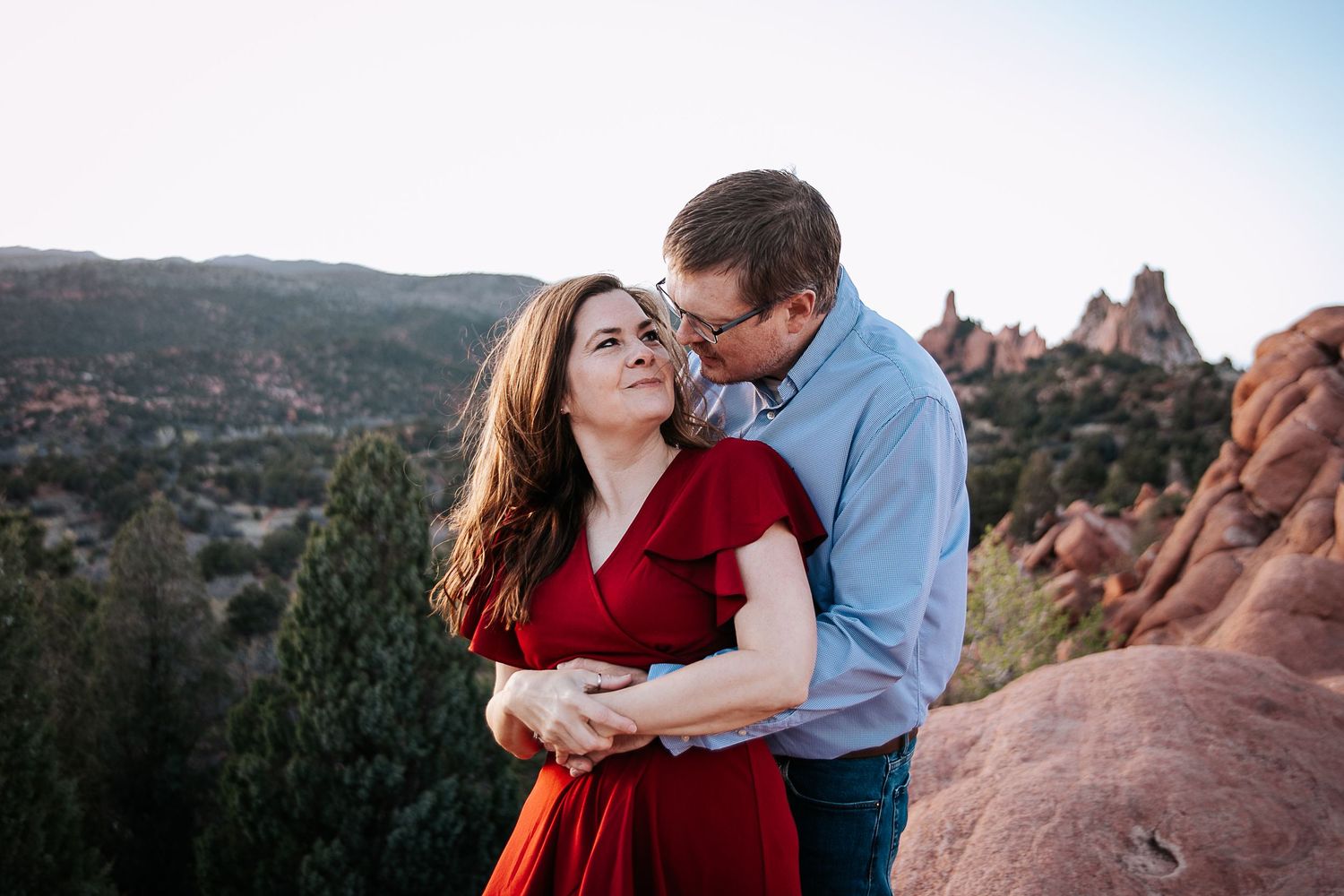 A couple embraces against a backdrop of scenic red rock formations and mountain vistas.
