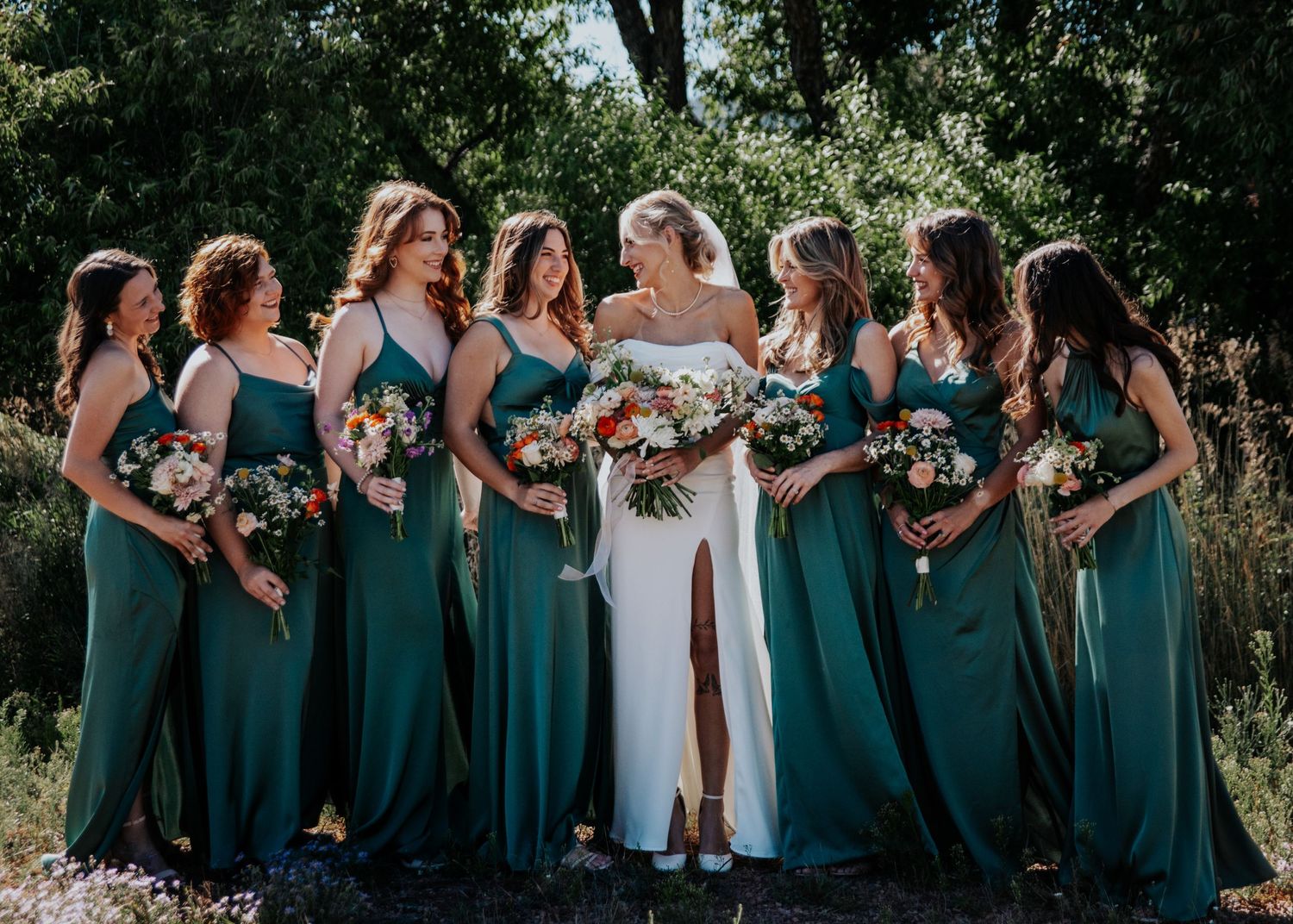 A bridal party in emerald green dresses poses together for a formal group photo outdoors.