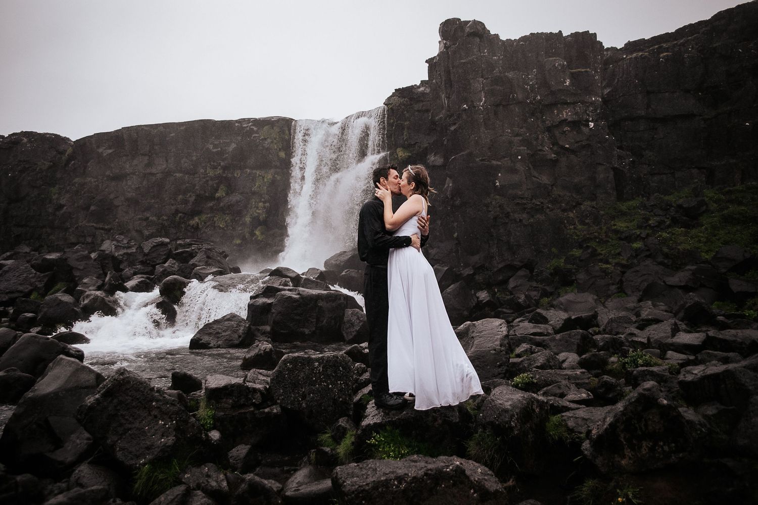 A couple shares an intimate moment against the dramatic backdrop of a cascading waterfall and dark rocky cliffs.