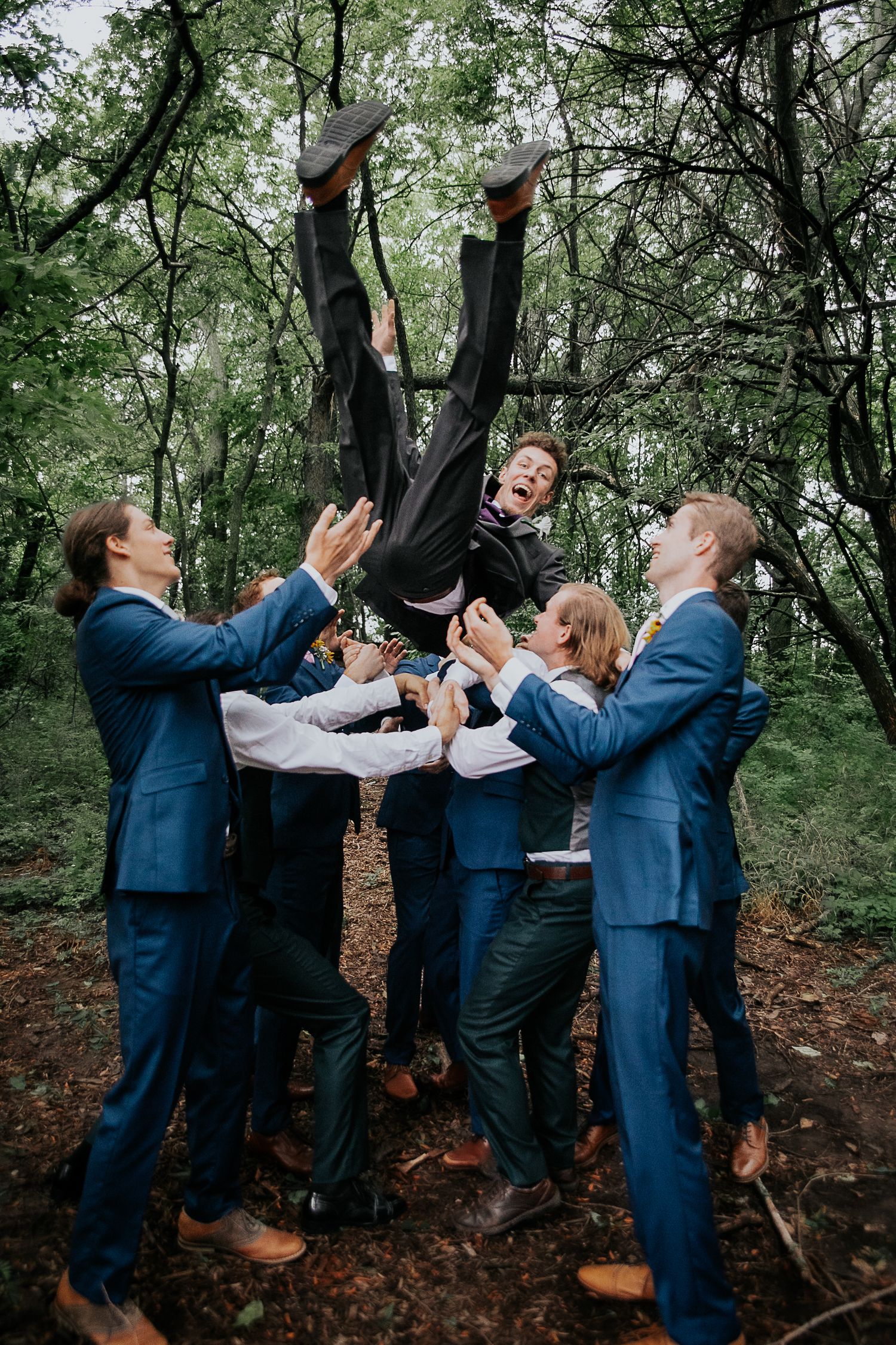 Wedding party in blue suits playfully posing in a forest setting.