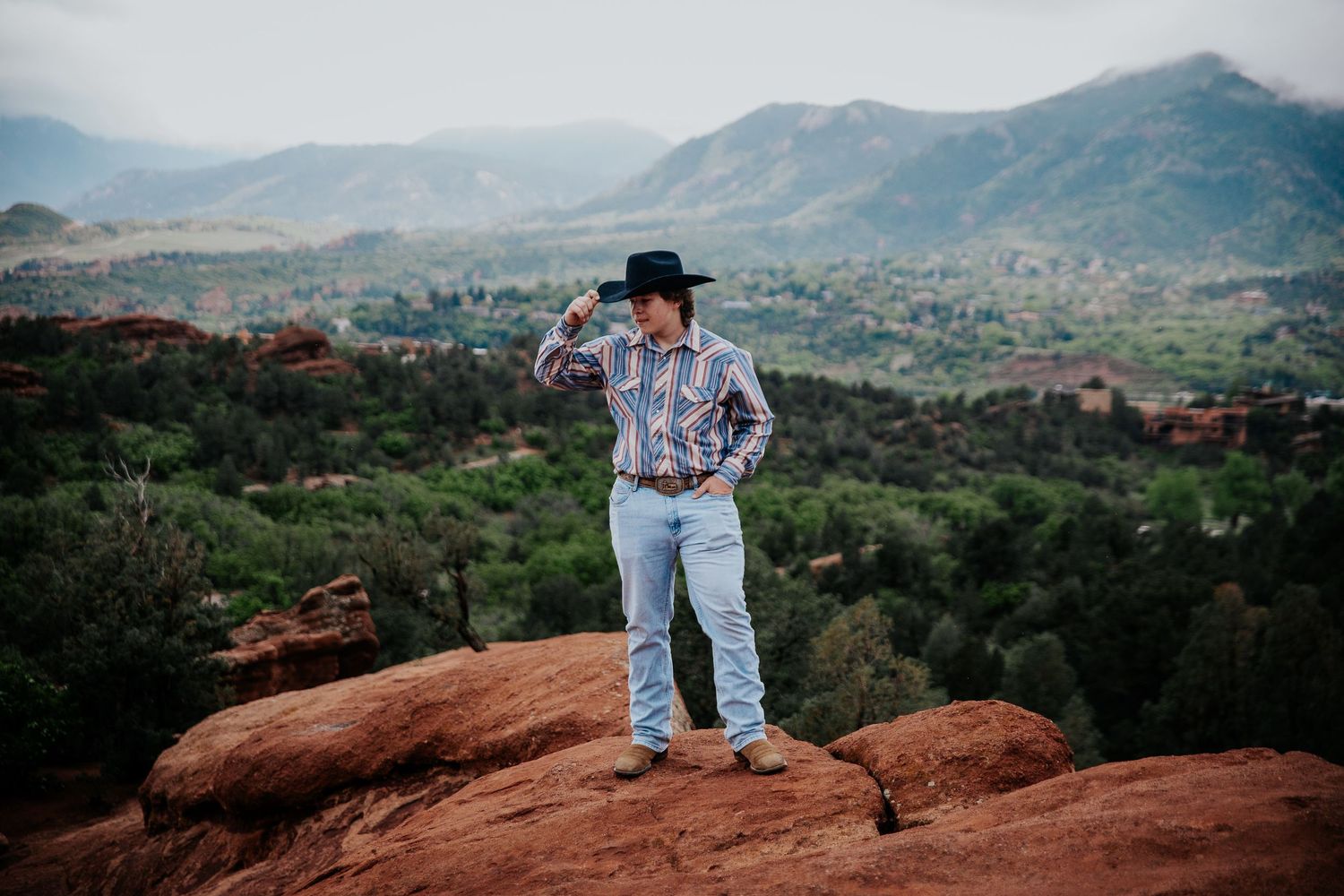 Person in western attire sits overlooking scenic mountain landscape from red rock formation at Garden of the Gods in Colorado Spri