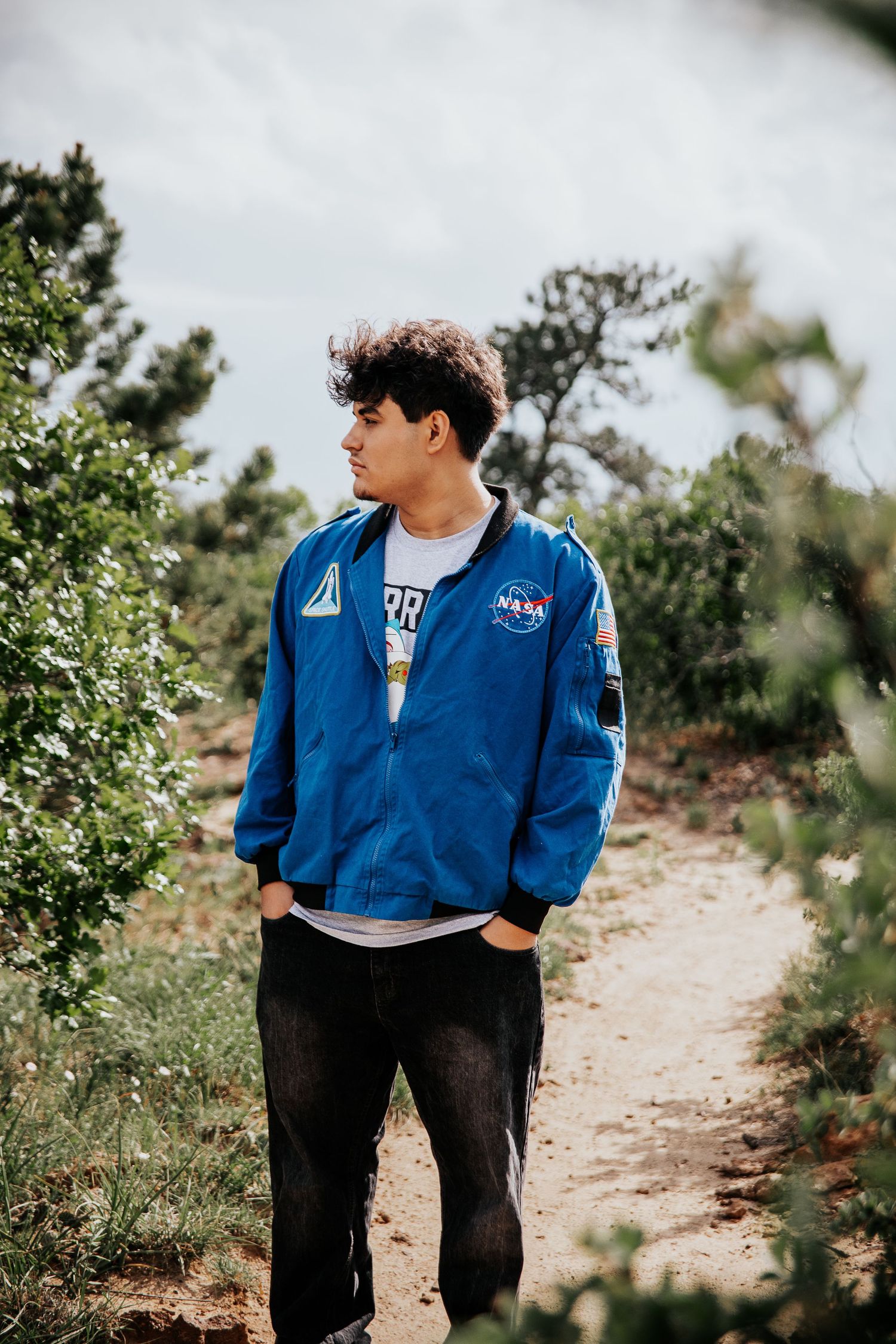 High school senior in NASA jacket stands on mountainside trail surrounded by greenery at Palmer Park in Colorado Springs