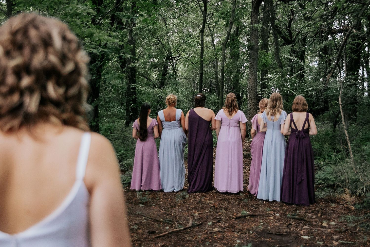Bridesmaids in varying shades of purple and pink dresses standing in a wooded area.