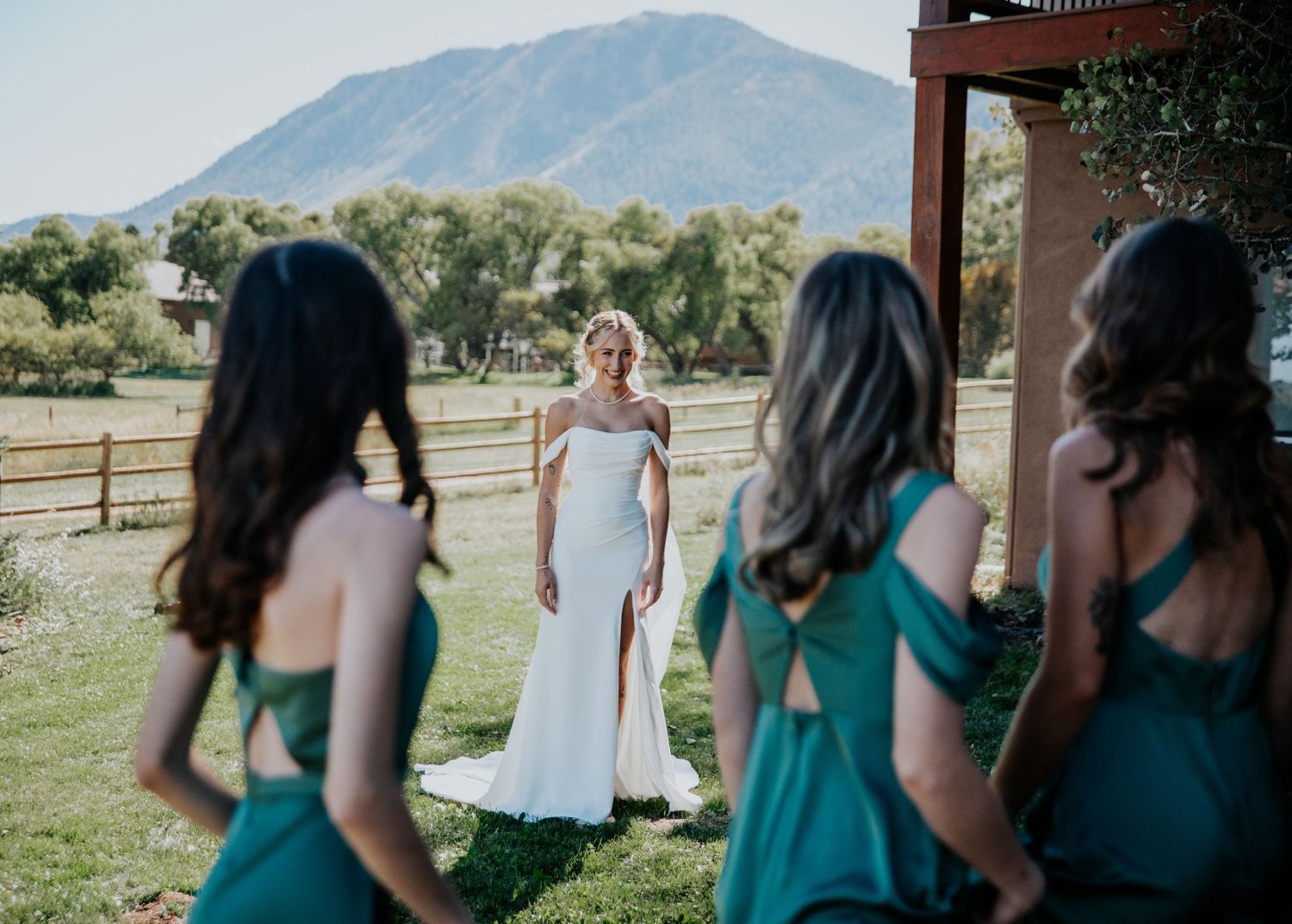 Bridesmaids in teal dresses admiring bride outdoors with mountain view.