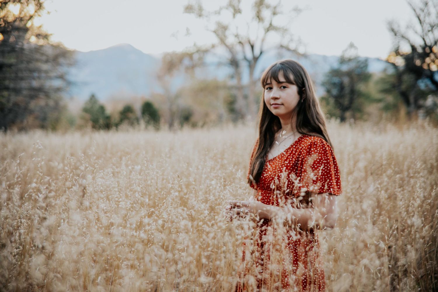 Woman in red dress poses in golden field with mountains visible in background.
