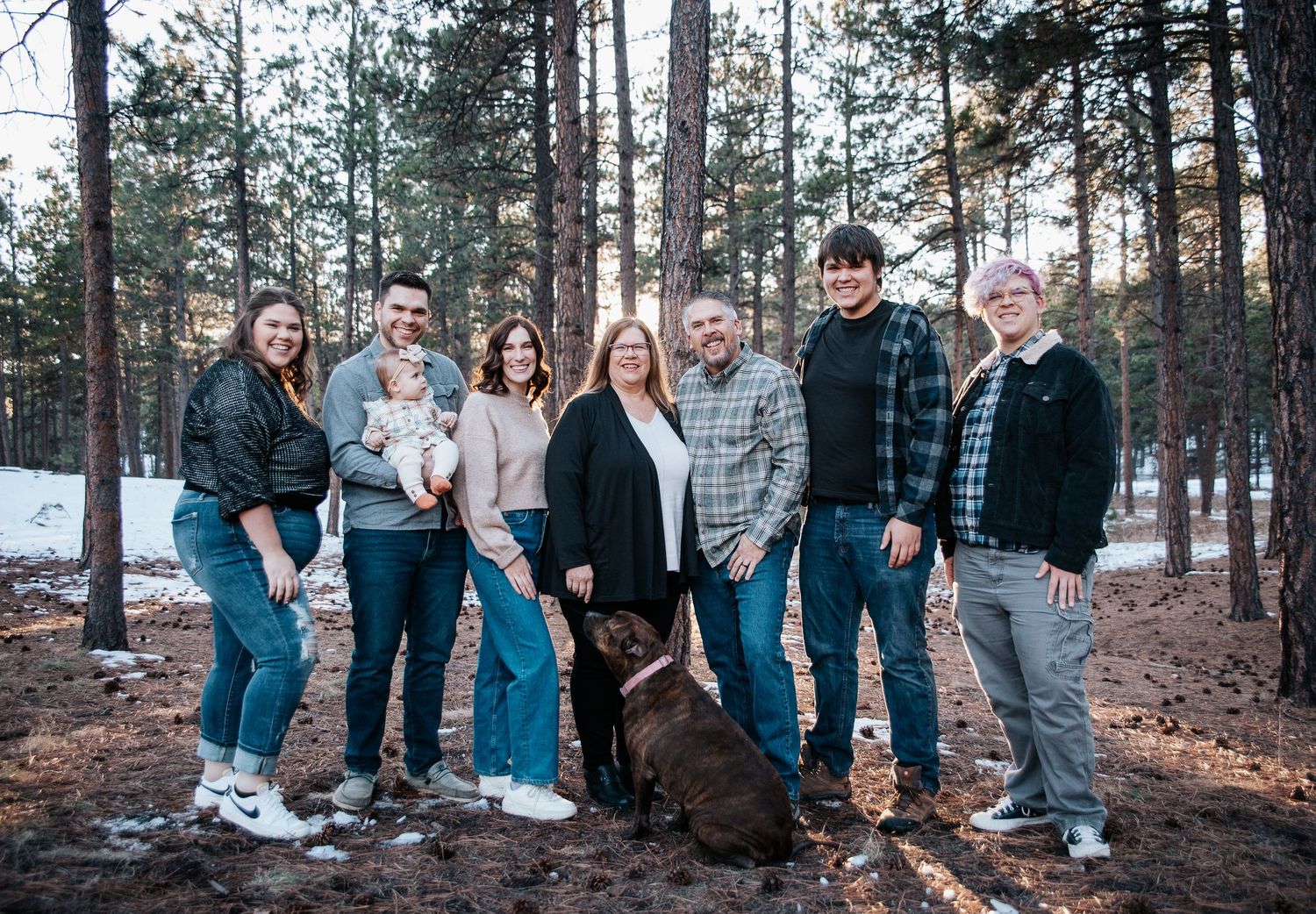 Friends and relatives stand together for a casual group photo among tall pine trees.
