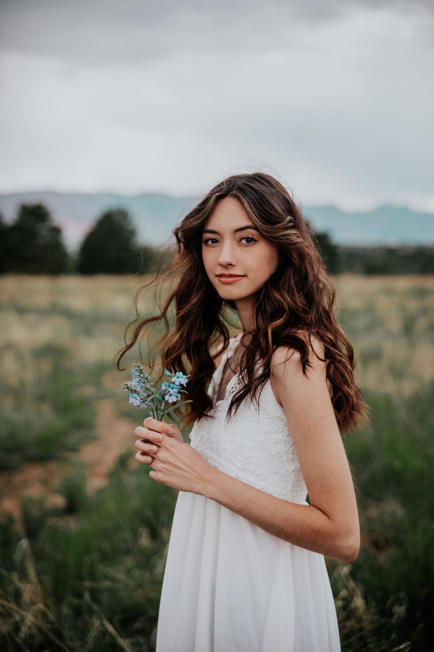 A person in white holds small blue flowers while posing in a natural field setting.