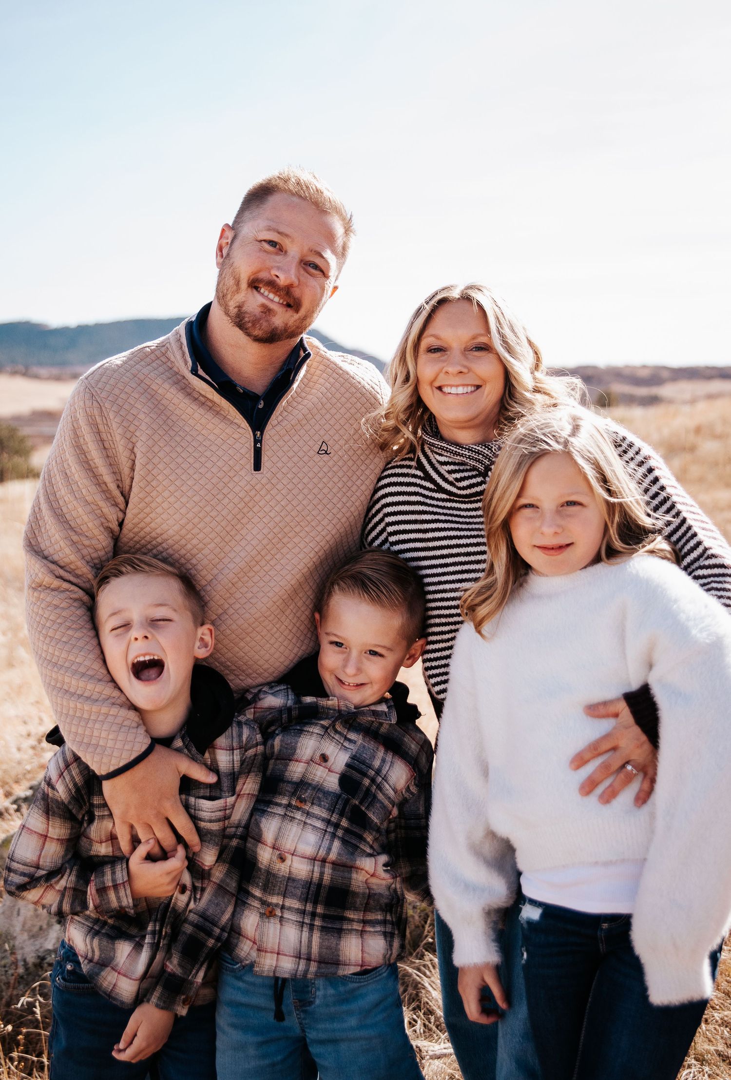 A family of four poses together outdoors in coordinating casual outfits with warm sweaters and plaid shirts.