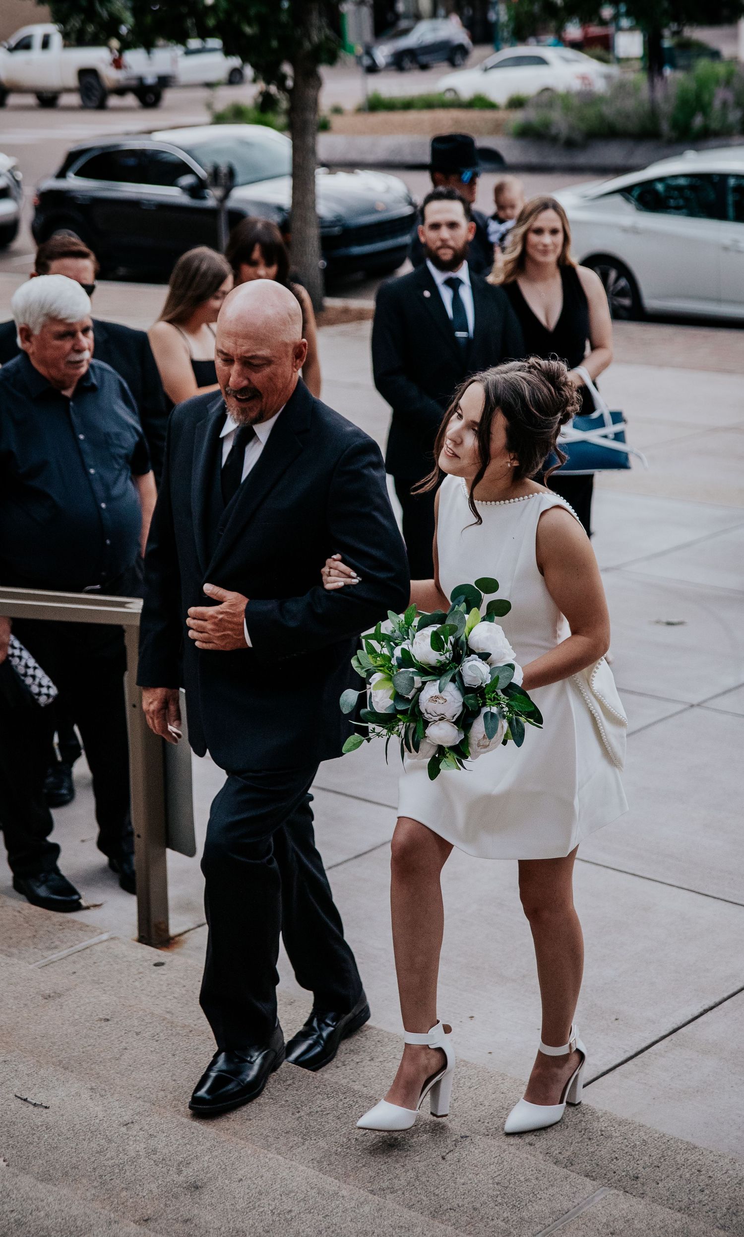Wedding party in formal attire gathering on sidewalk outside urban venue.