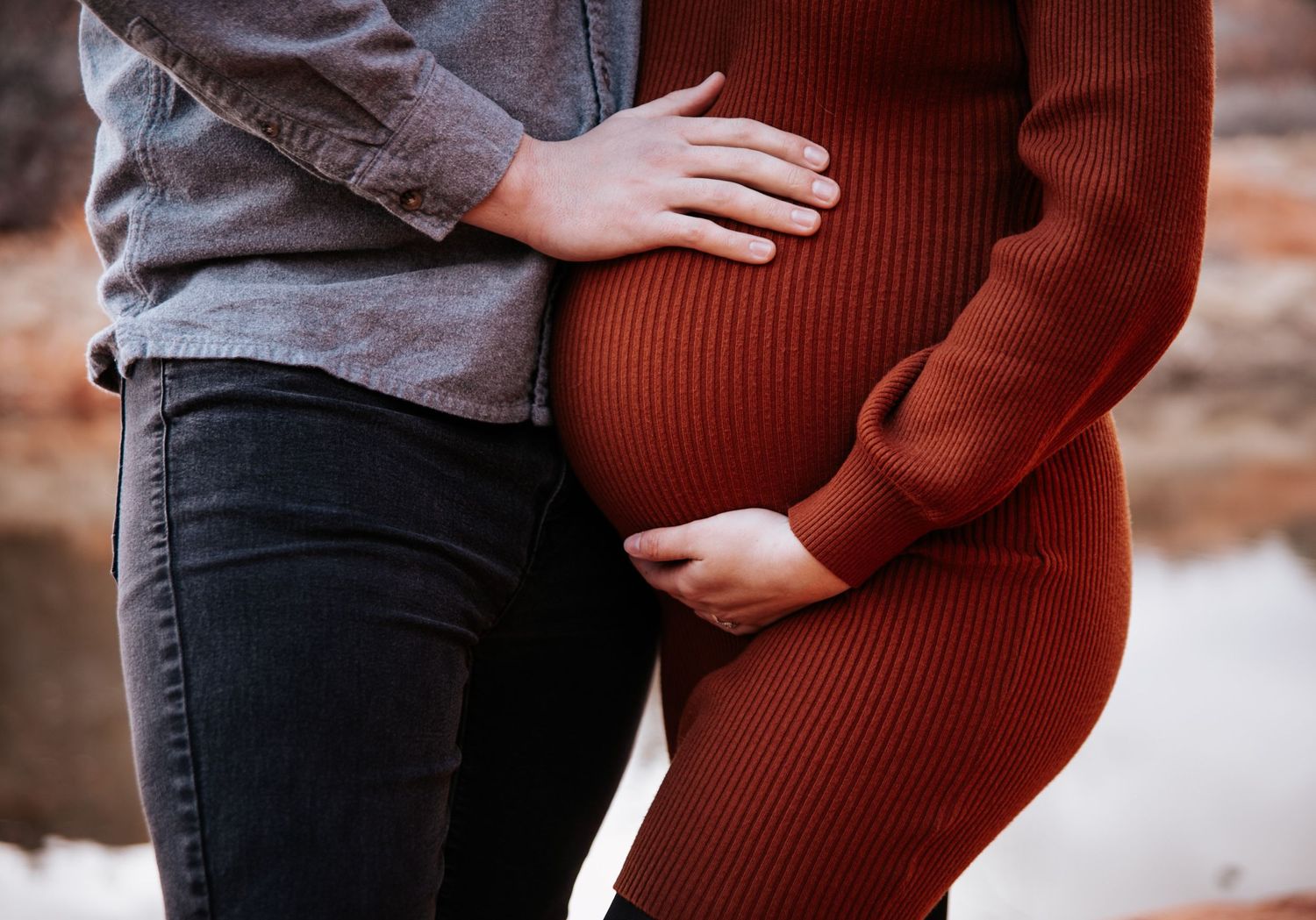 Close-up shot of a pregnant woman in a burgundy sweater dress with her partner's hands on her belly.