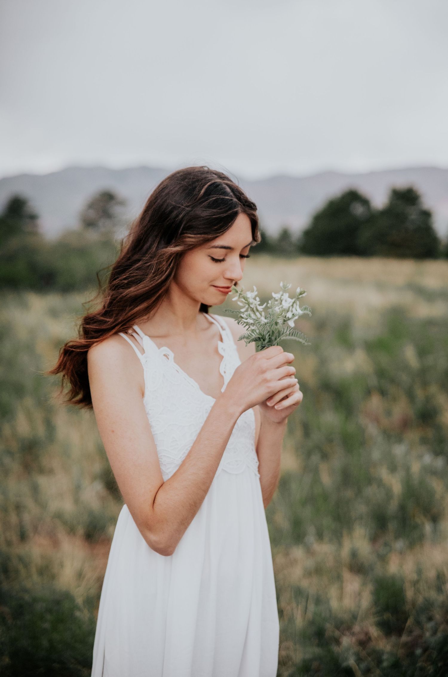 A person in white holding wildflowers in a meadow with mountains behind.