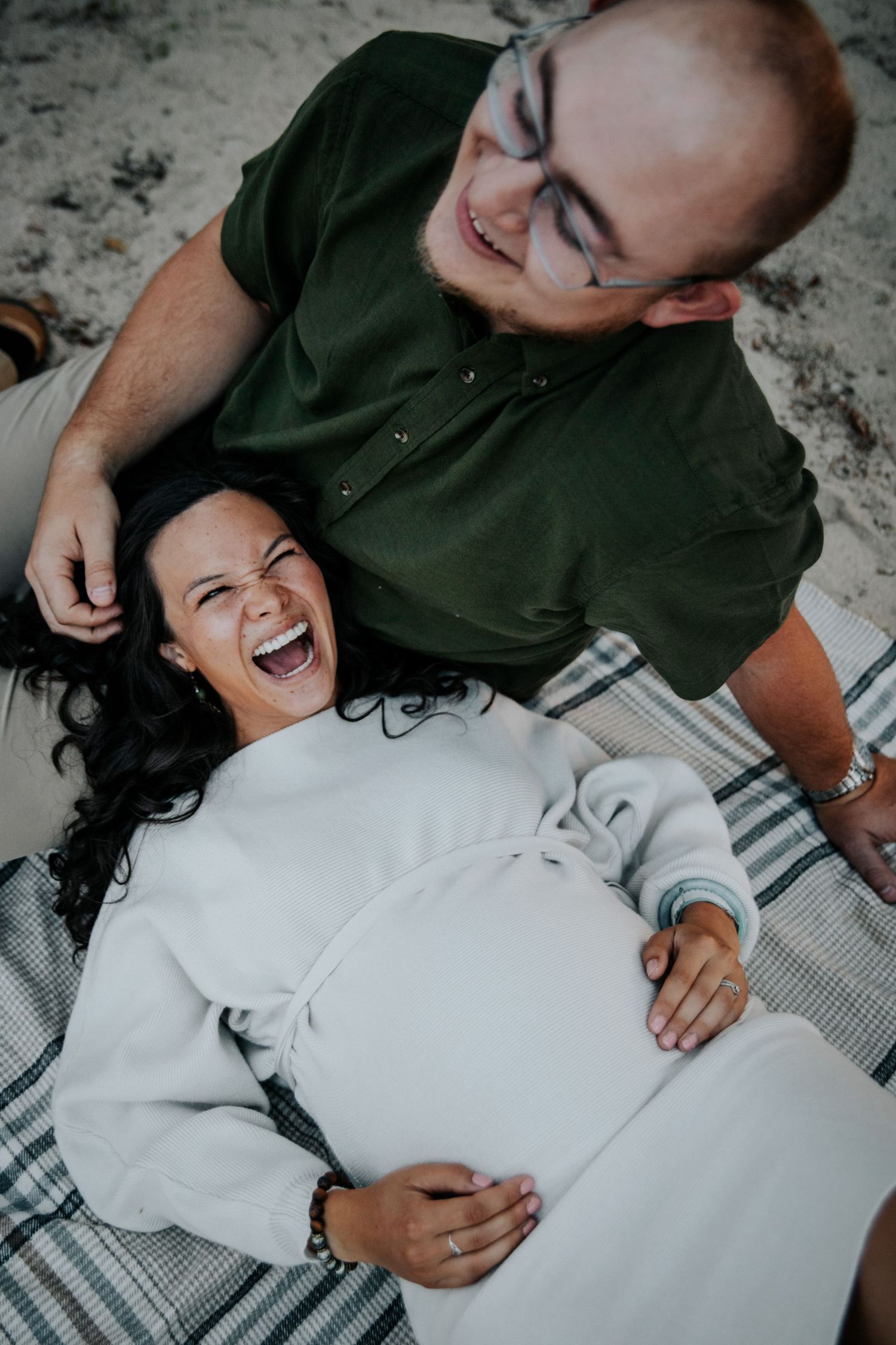 A couple shares joyful laughter while lying on a plaid blanket outdoors.