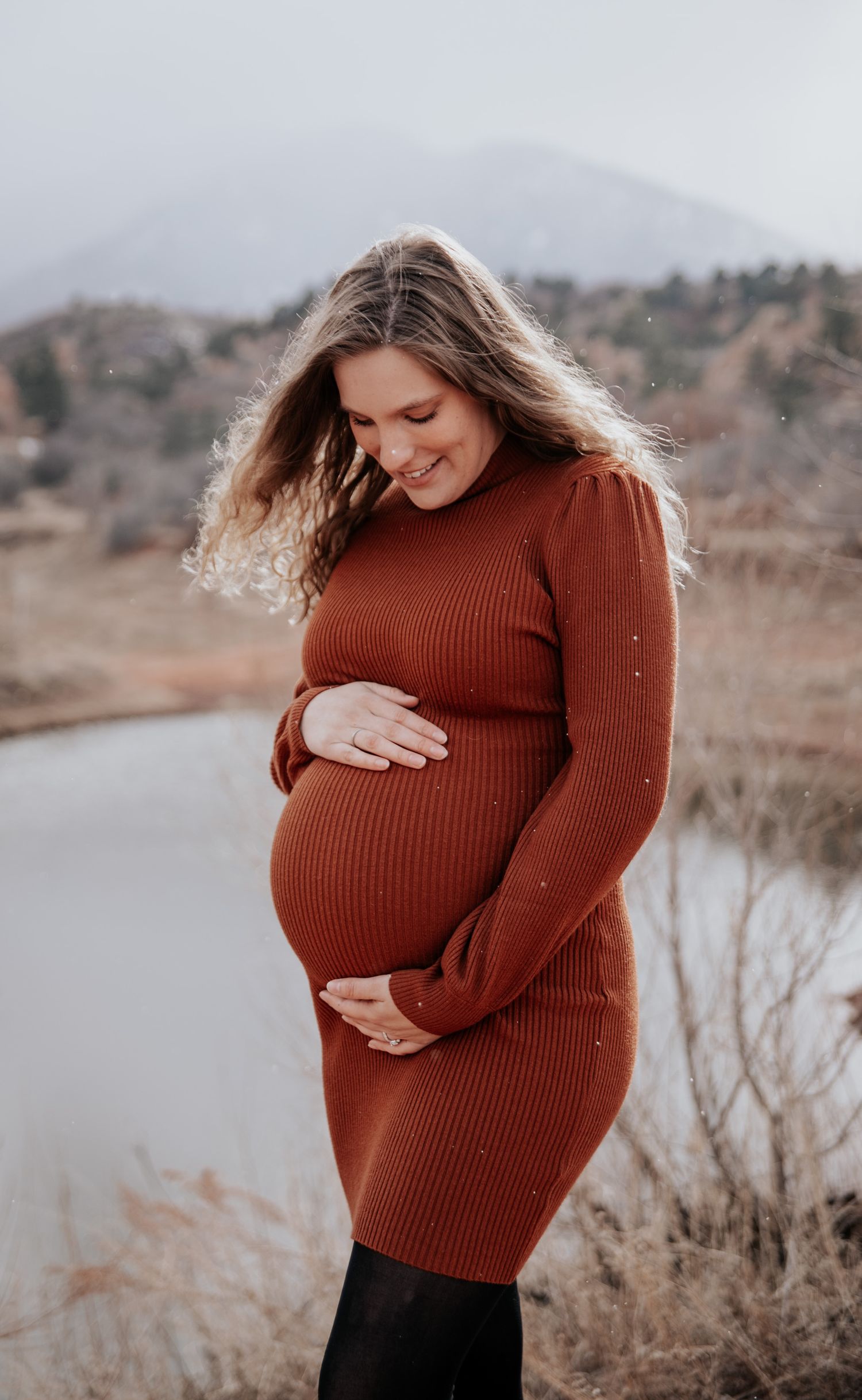 A person in a ribbed rust sweater cradles their baby bump against a misty mountain backdrop.