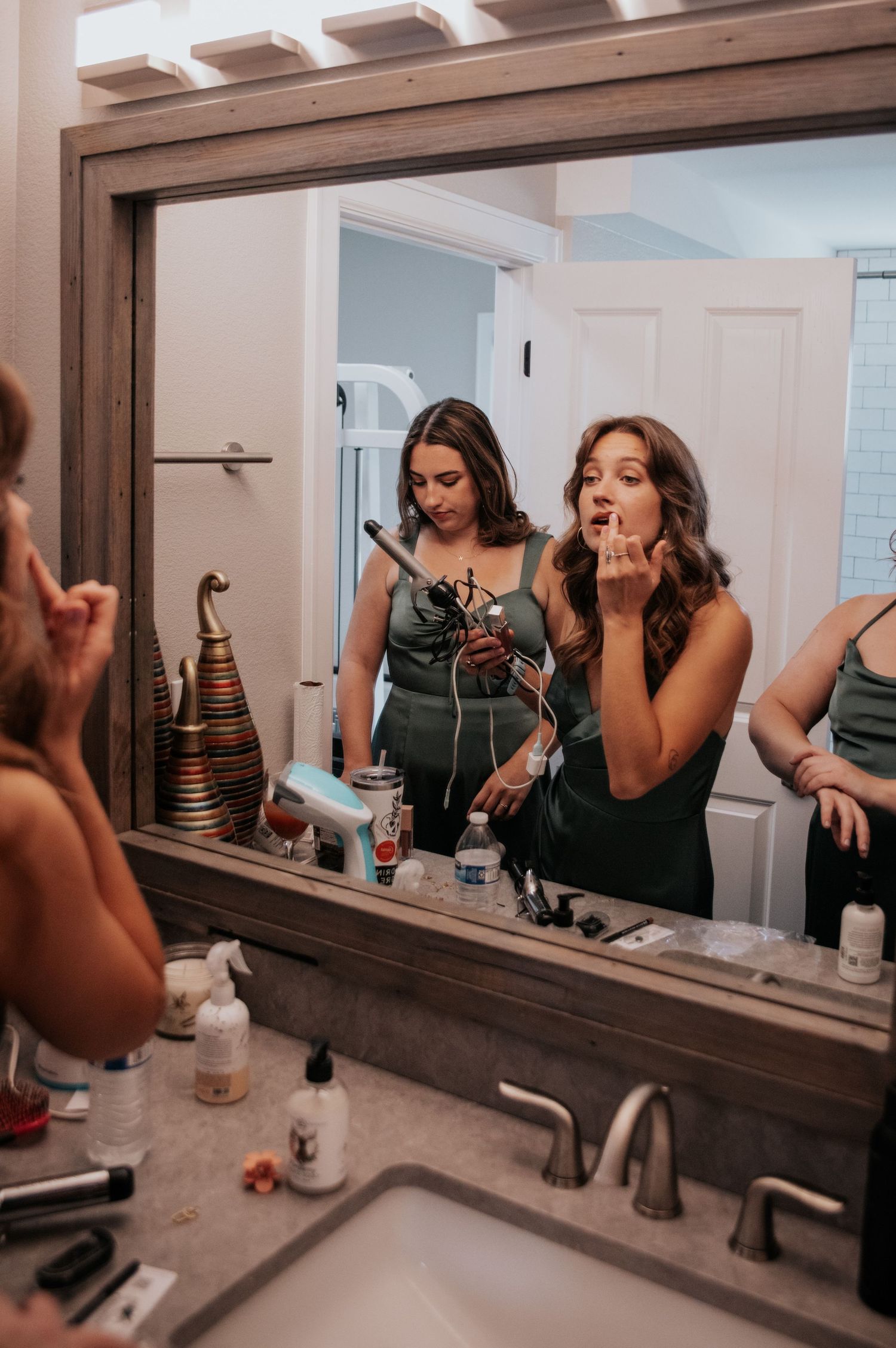 Two bridesmaids in emerald green dresses doing makeup in a bathroom mirror.