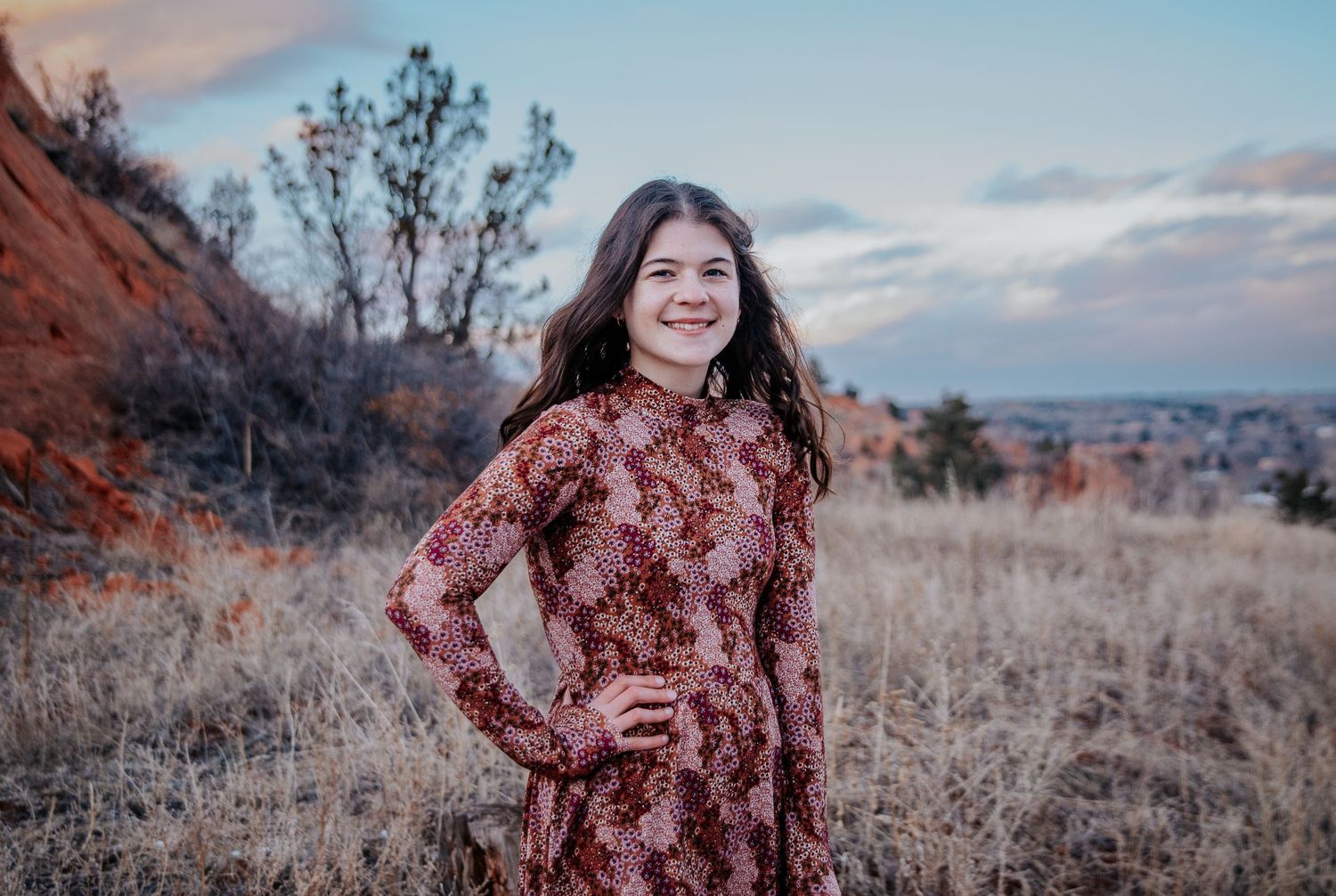 A woman in a burgundy dress poses in front of a scenic desert landscape at sunset.