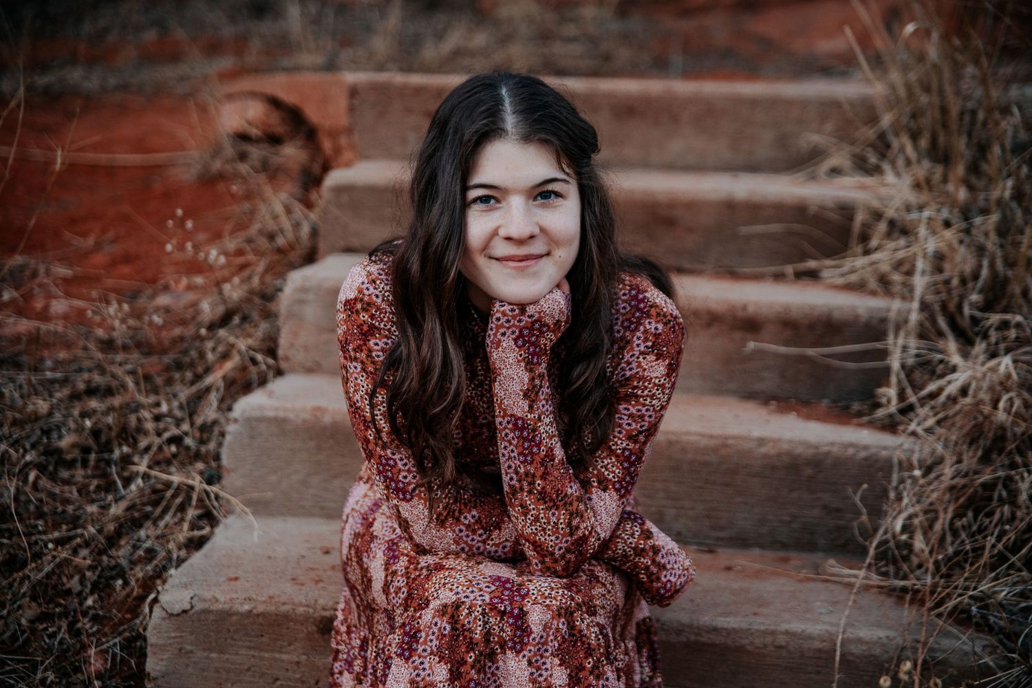 Portrait of a woman in a patterned dress sitting on red stone steps outdoors.