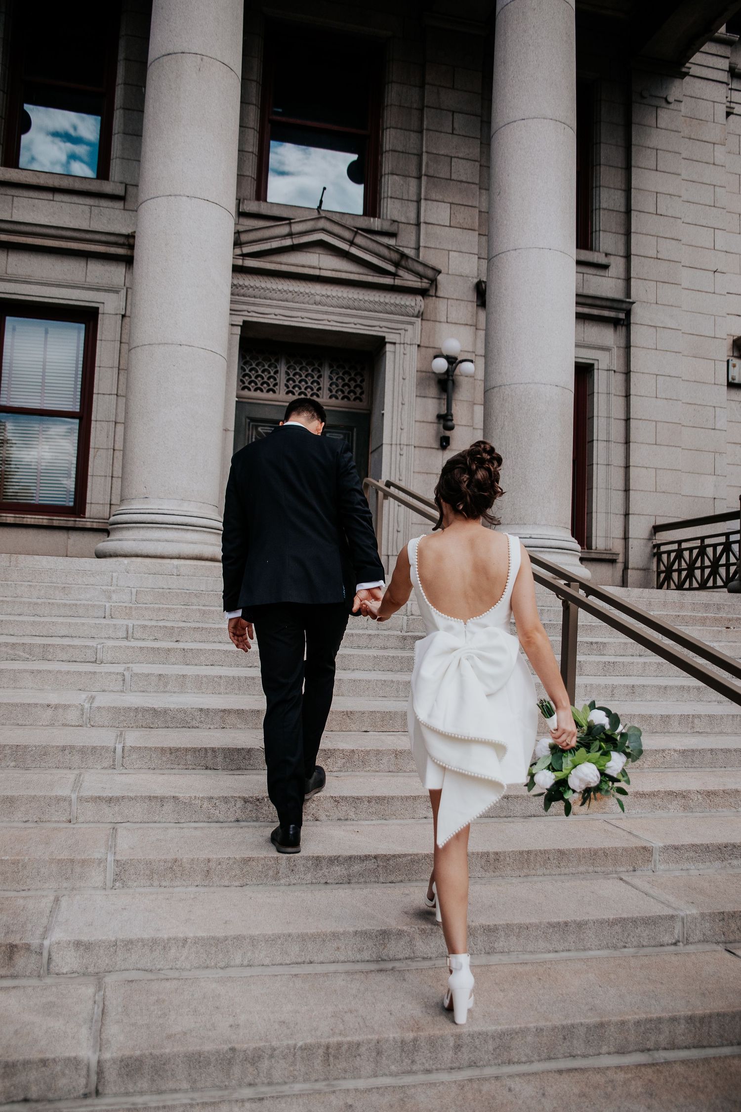 A wedding couple ascends stone steps of a grand building, with the bride's white dress and bouquet visible from behind.
