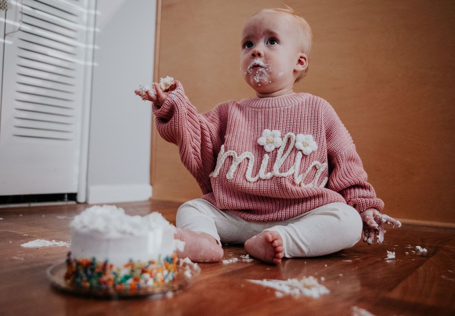 A pink-sweater-clad baby explores cake frosting during first birthday celebration.