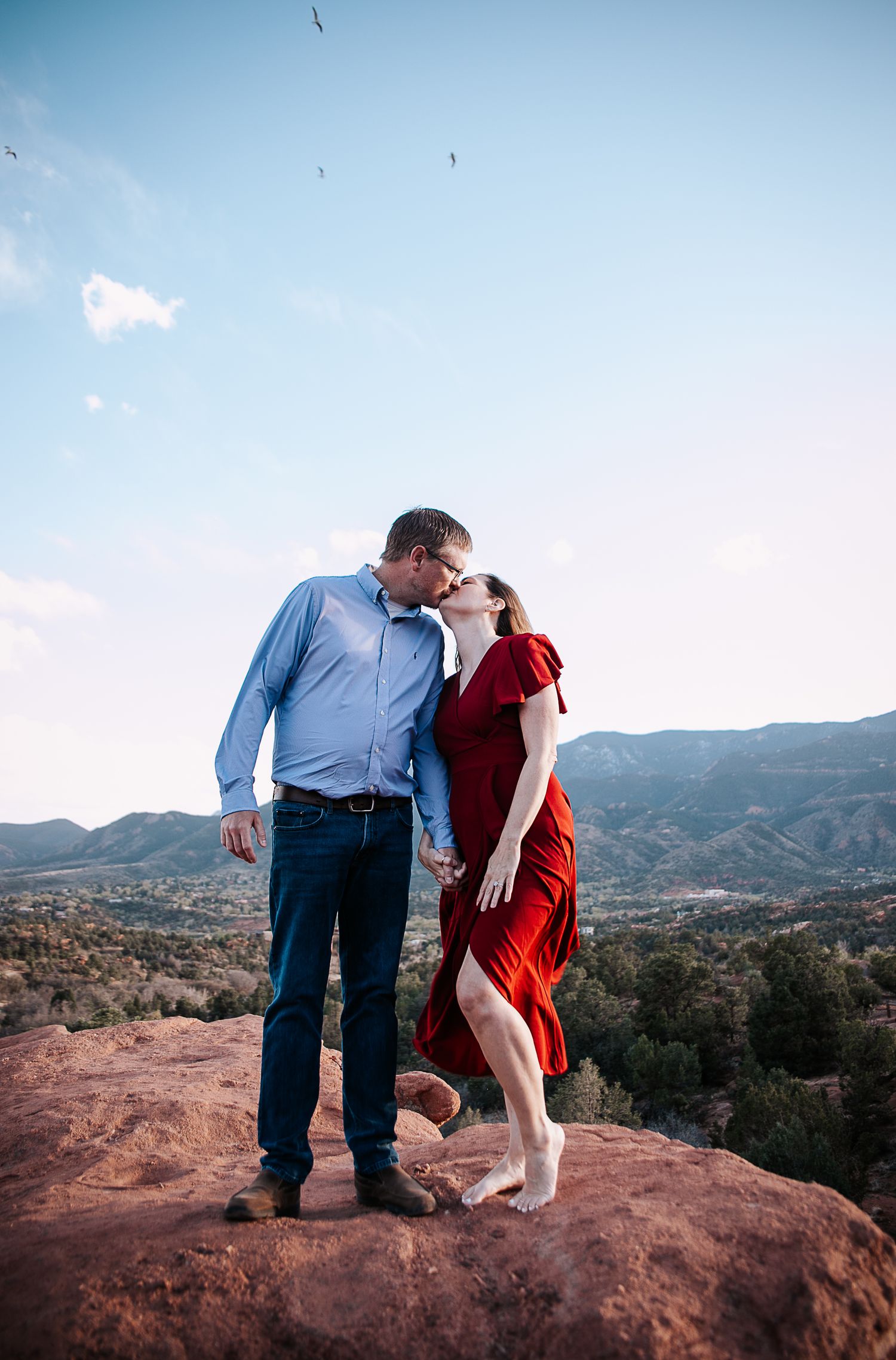 A romantic couple shares a tender moment on red rocks overlooking a mountain landscape at sunset.
