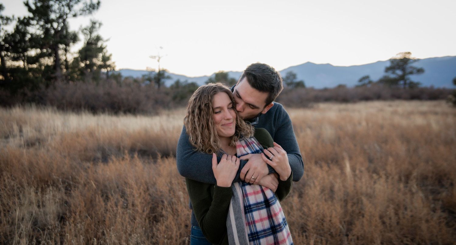 Intimate embrace between couple in field with mountains in background at sunset.