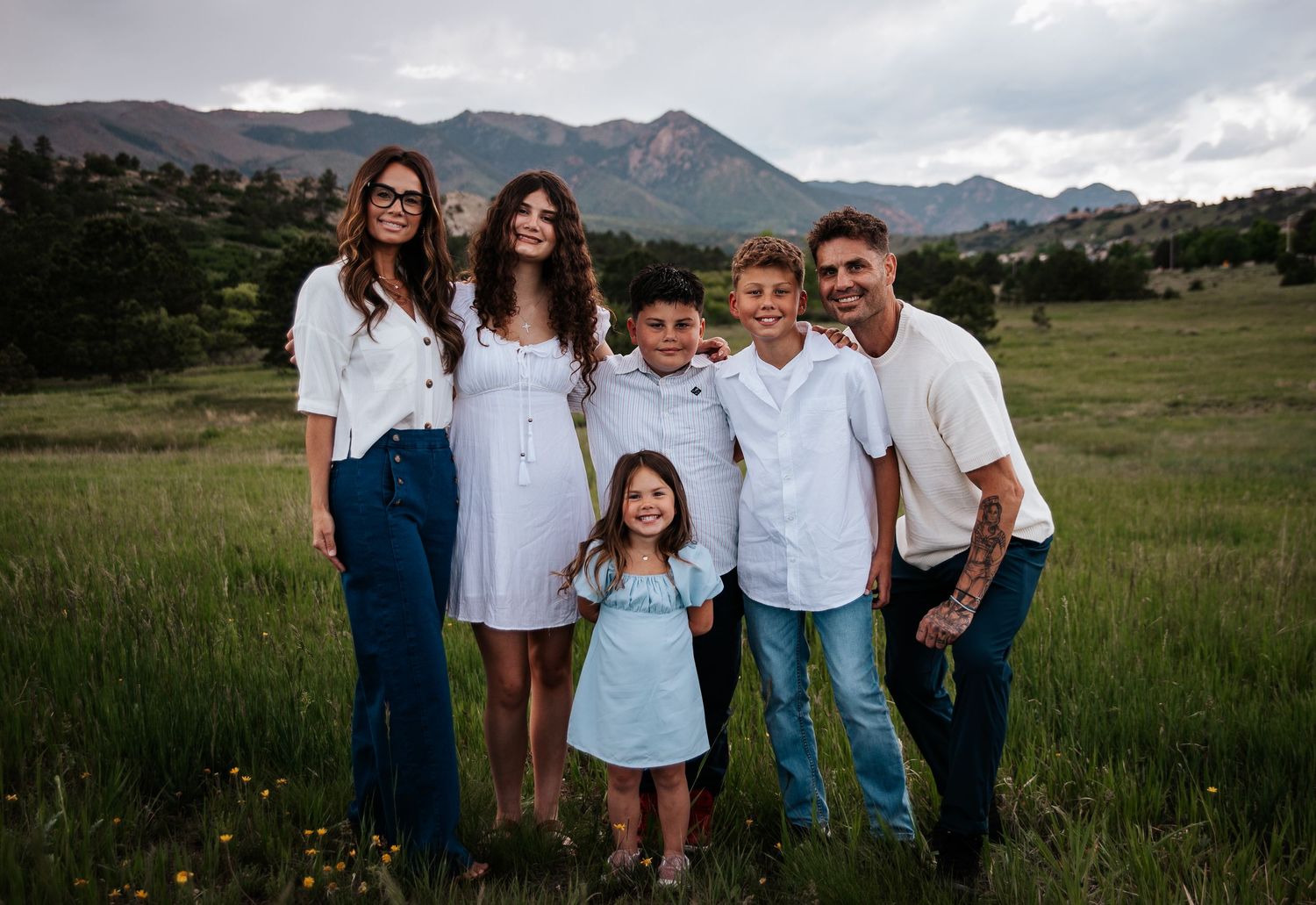 A family dressed in white and blue poses together in a grassy mountain meadow at sunset at Ute Valley Park in Colorado Springs