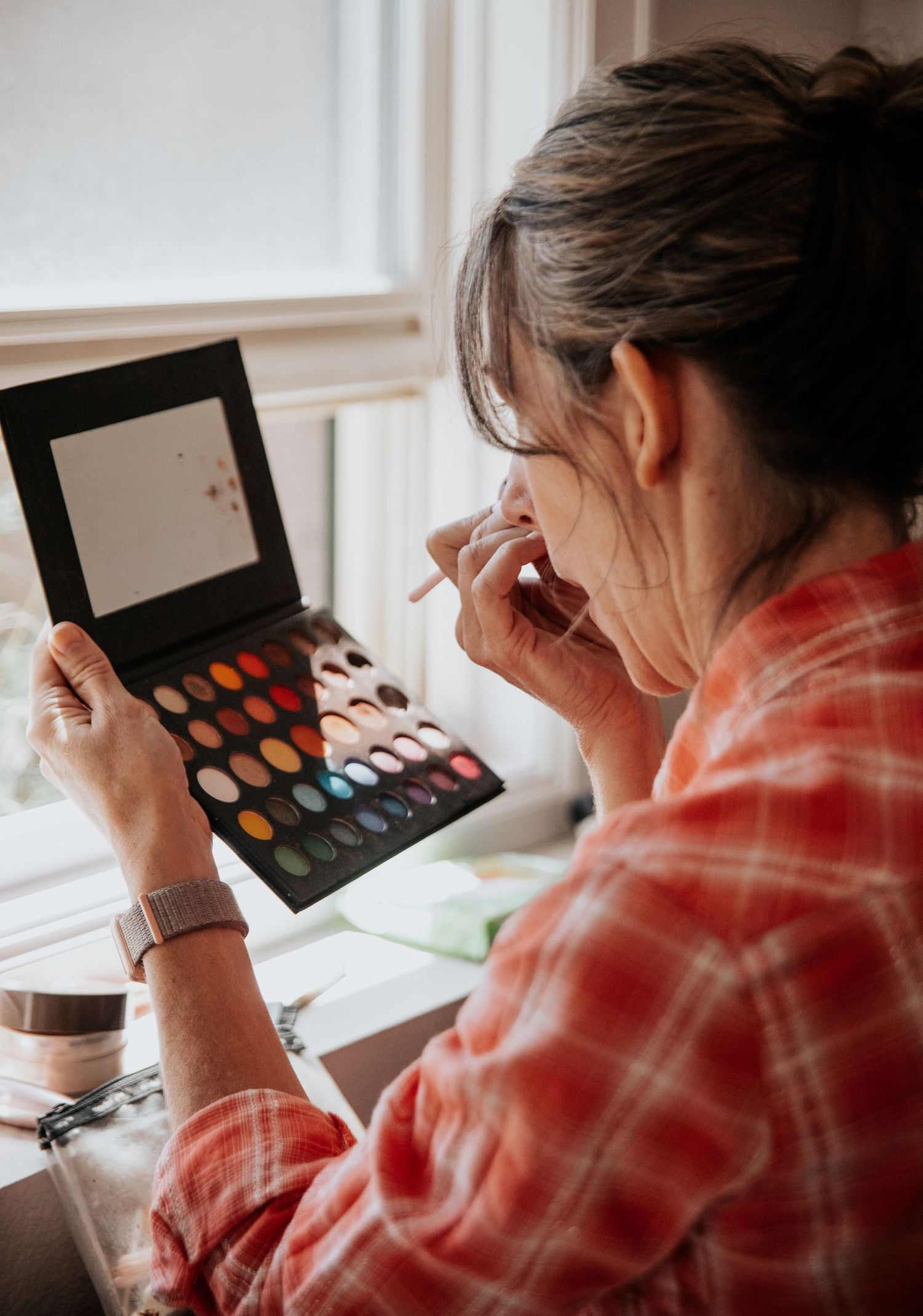 Person in plaid shirt looking at makeup palette near window in soft natural light.
