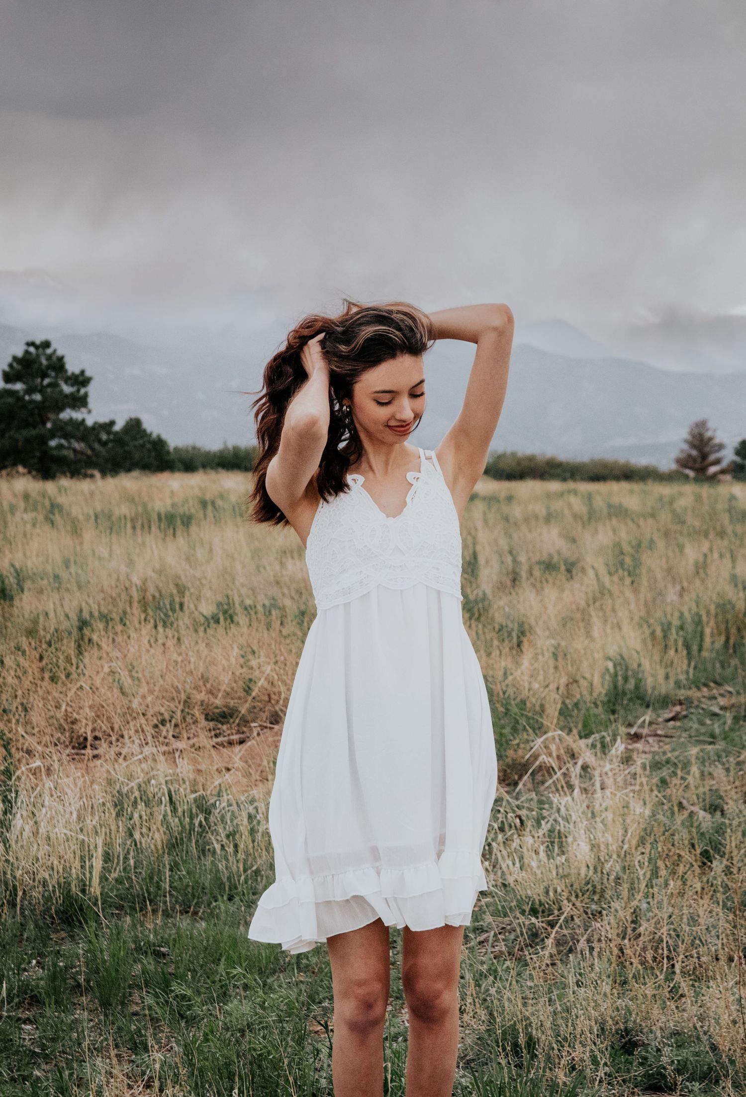 Woman in white flowing dress twirls playfully in a grassy field with mountains in the background.