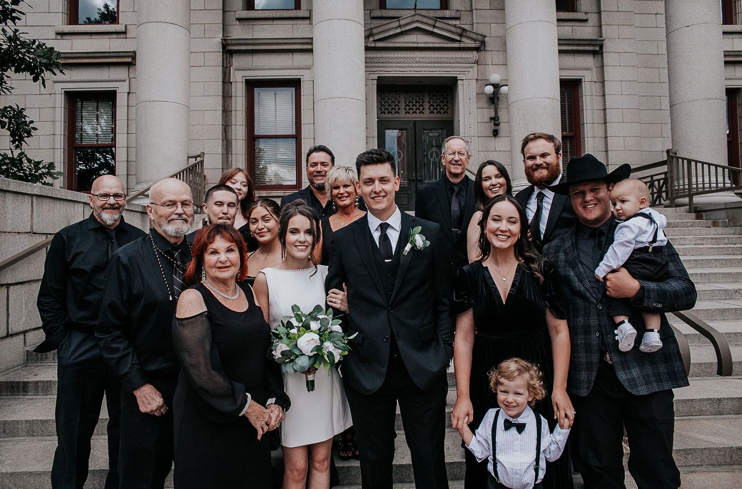 Wedding party dressed in black and white formal attire poses on stone steps of historic building.